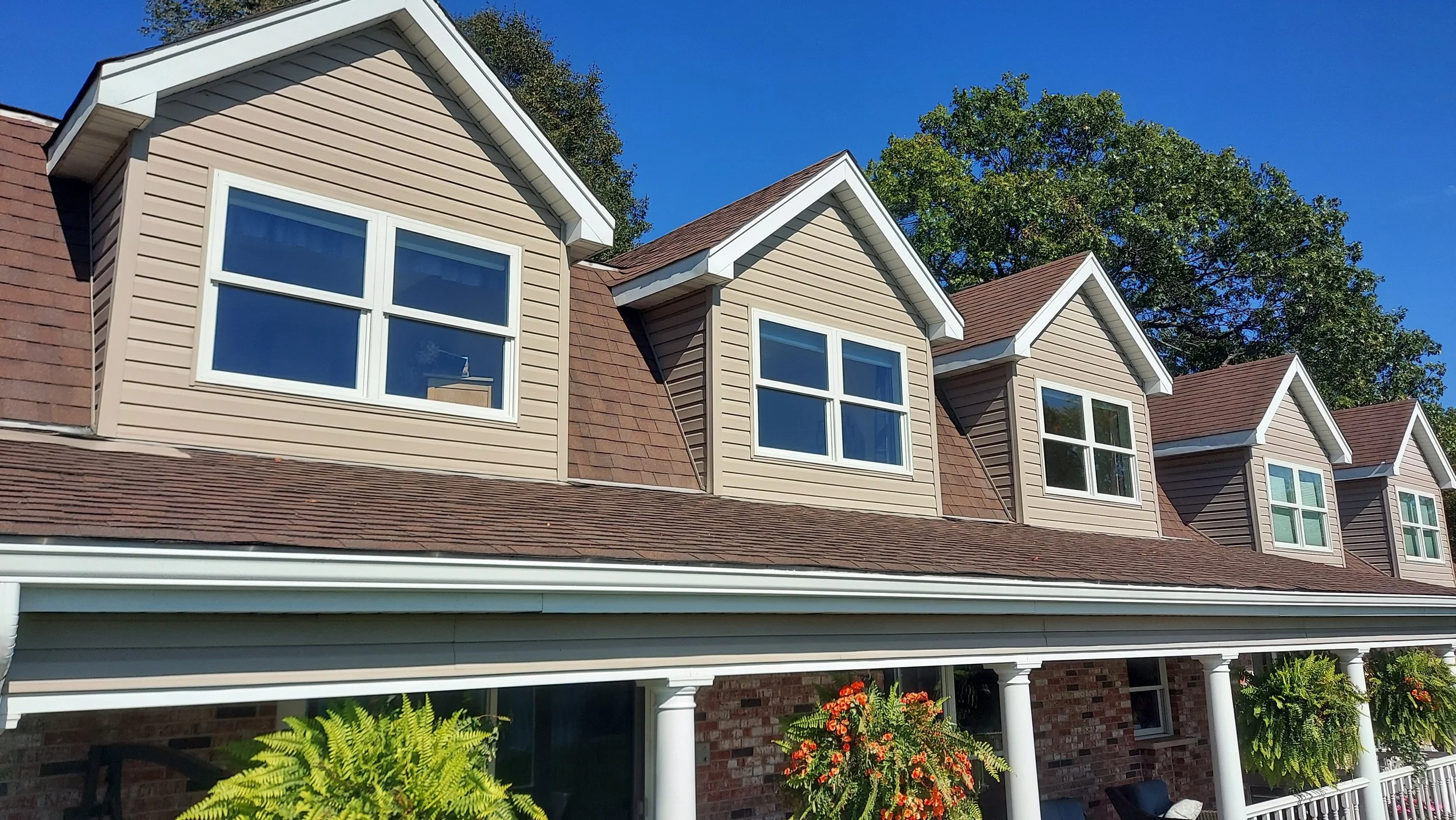 Row of townhouses with beige siding, brown shingle roofs, and white-framed windows, with a porch featuring white columns and hanging plants, against a blue sky with trees in the background.
