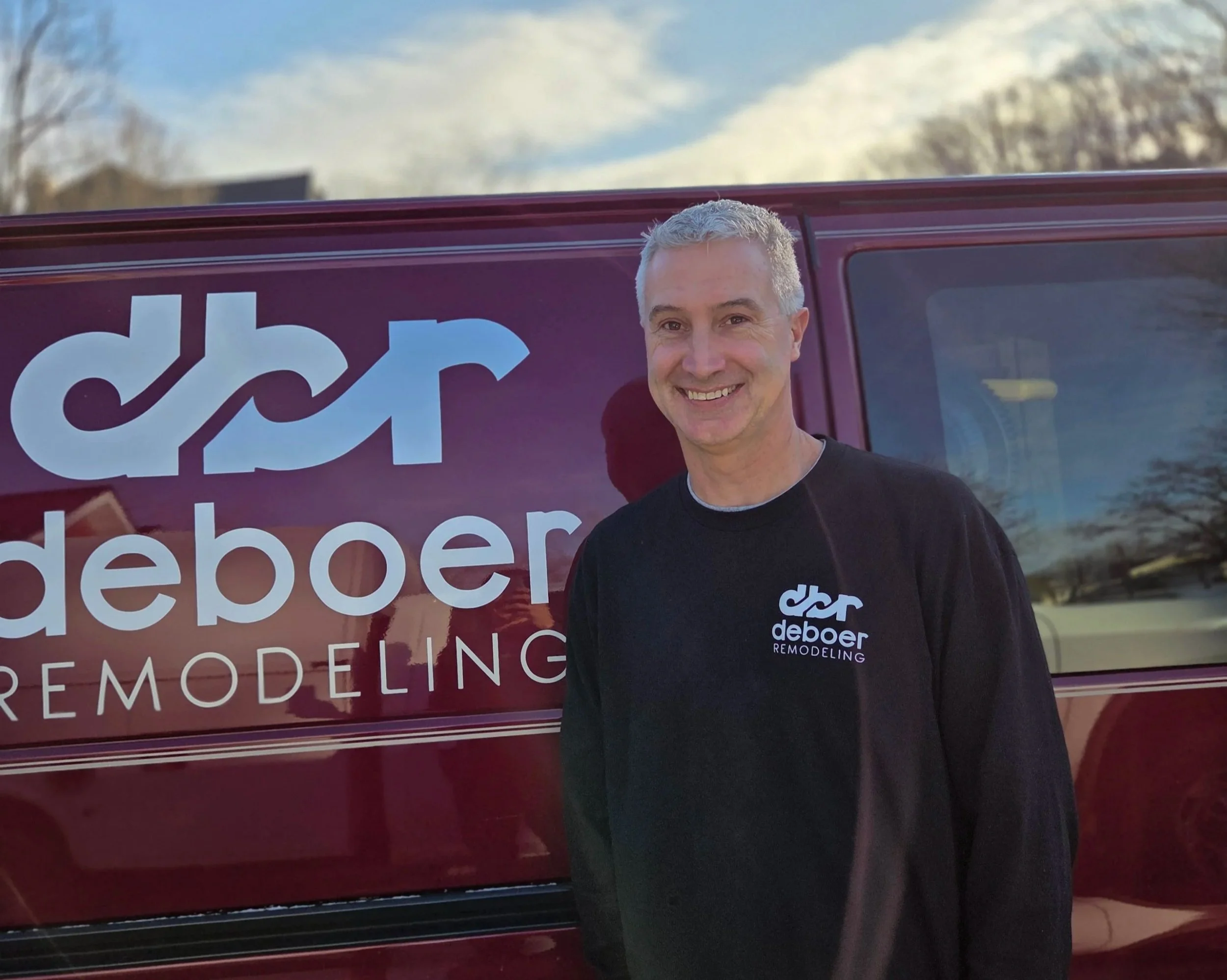 A smiling man with short gray hair wearing a black 'd&r deboer REMODELING' shirt standing in front of a maroon company van with the same logo, parked outdoors on a clear day with trees and a blue sky in the background.