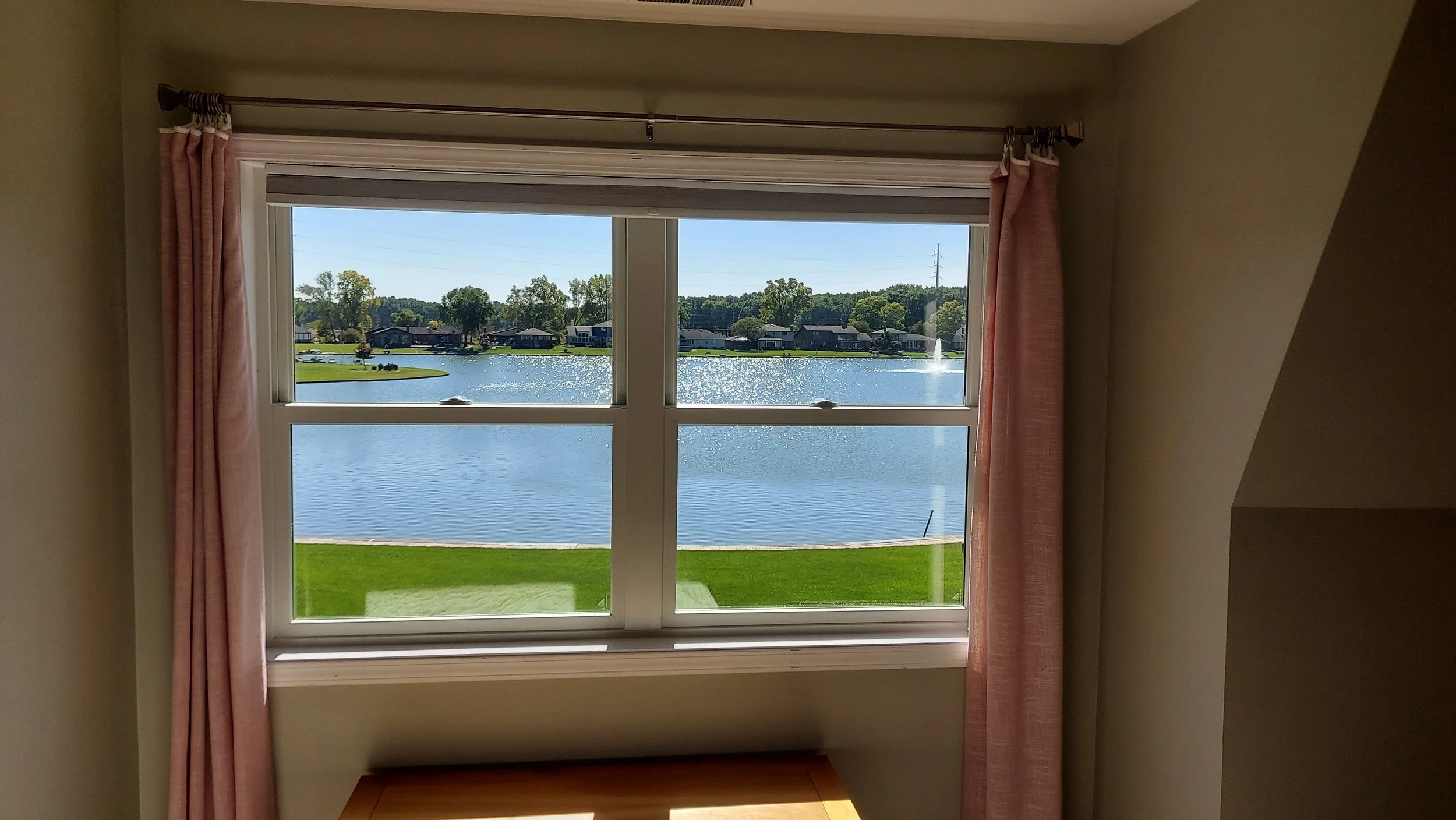 View through a window showing a lake, green grass, houses, and a fountain on a sunny day.