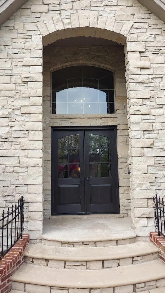 Entryway of a house with a black double door, stone arch, and brick steps with black metal railings.