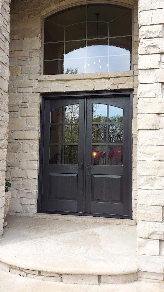 Double black doors with glass panes and a semi-circular transom window above, set in a stone facade entrance with a small concrete step.