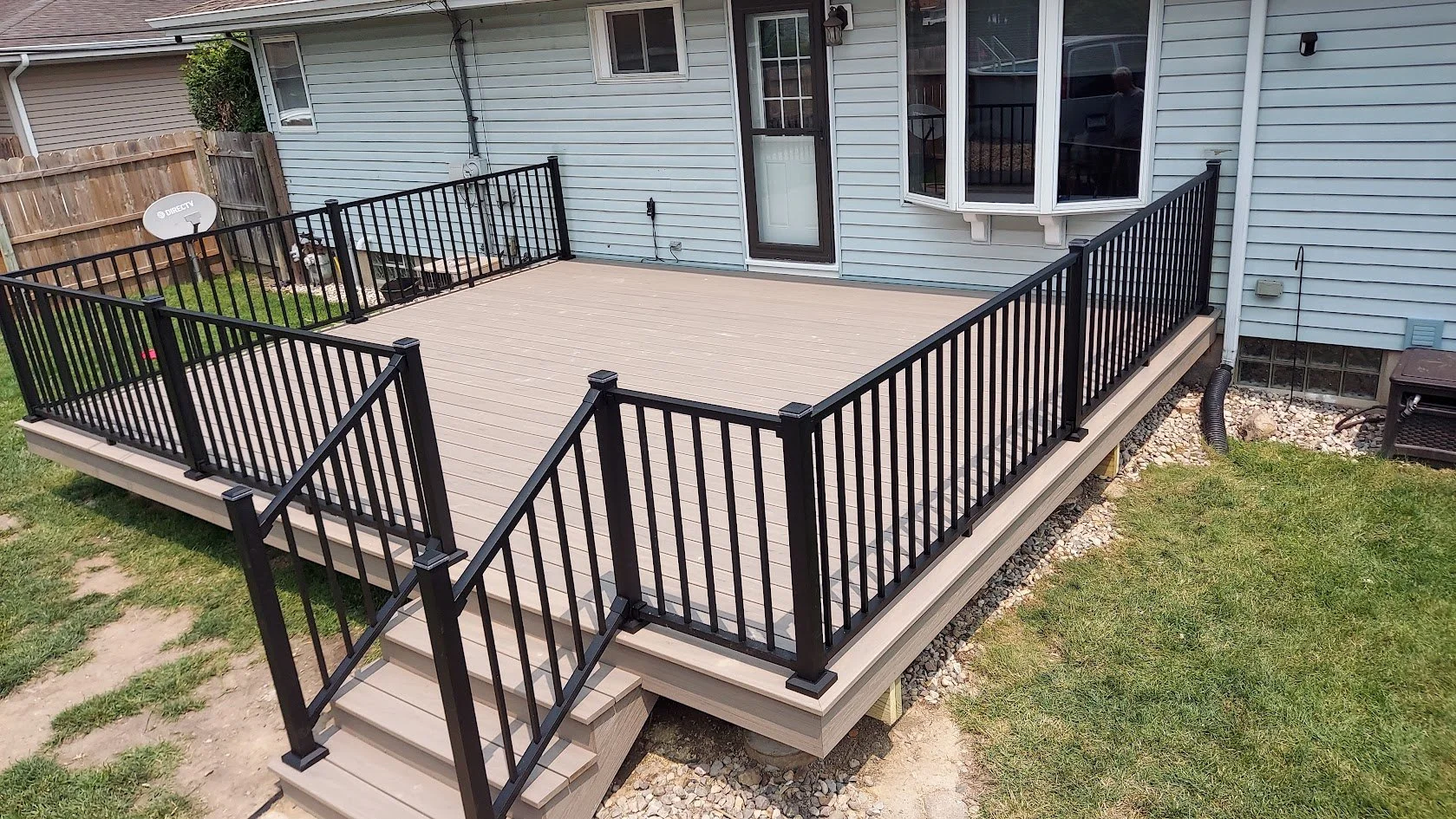Newly built elevated wooden deck with black metal railing outside a house, accessed by stairs, with a small lawn and gravel area underneath.