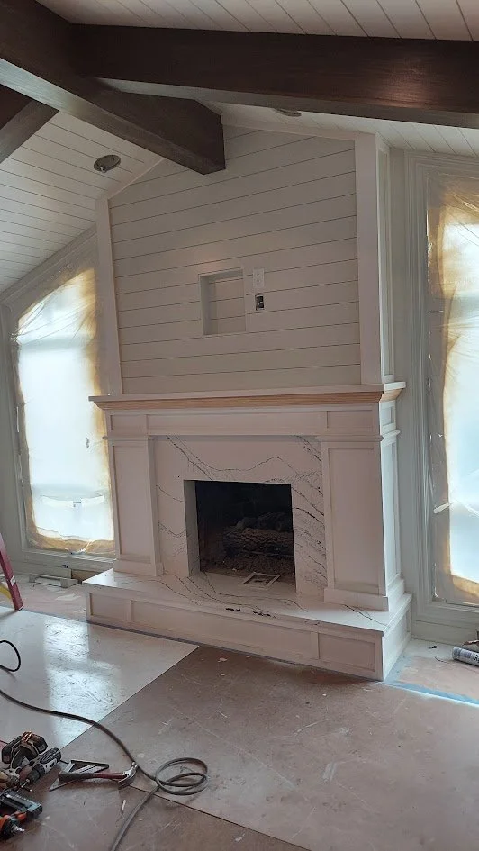 Living room with a white marble fireplace on a wooden mantle, flanked by two large windows covered with sheer yellow curtains, inside a house under renovation.