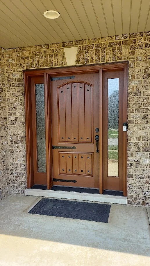 A wooden front door with black hardware, flanked by sidelights with frosted glass, set into a brick exterior wall under a beige ceiling. There is a black welcome mat on a concrete porch in front of the door.