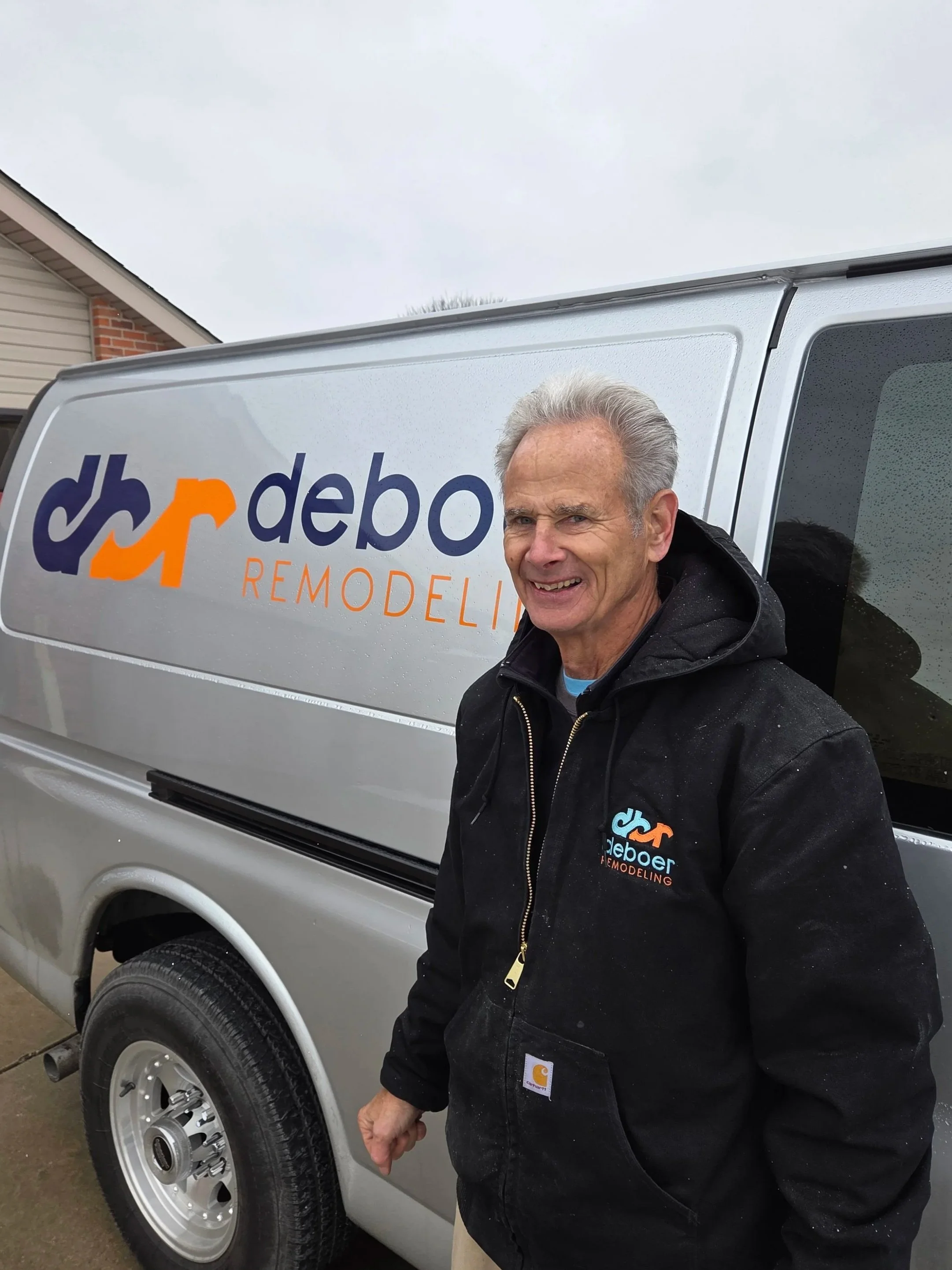 A smiling man with gray hair standing next to a silver remodeling company's van. The van has the logo 'djr redecor' on the side, with the words 'REMODELING' underneath.