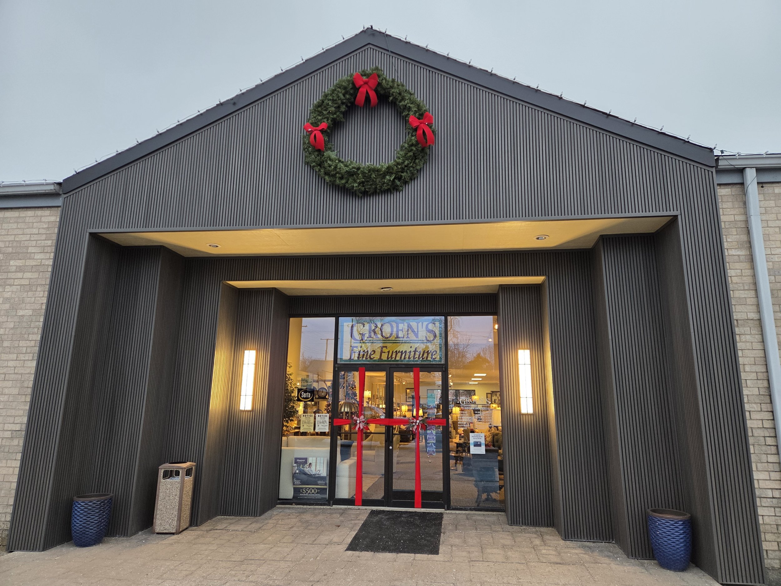 Store entrance decorated with Christmas wreath and red bows, with illuminated wall lights and glass doors, indoors furniture visible through the glass.