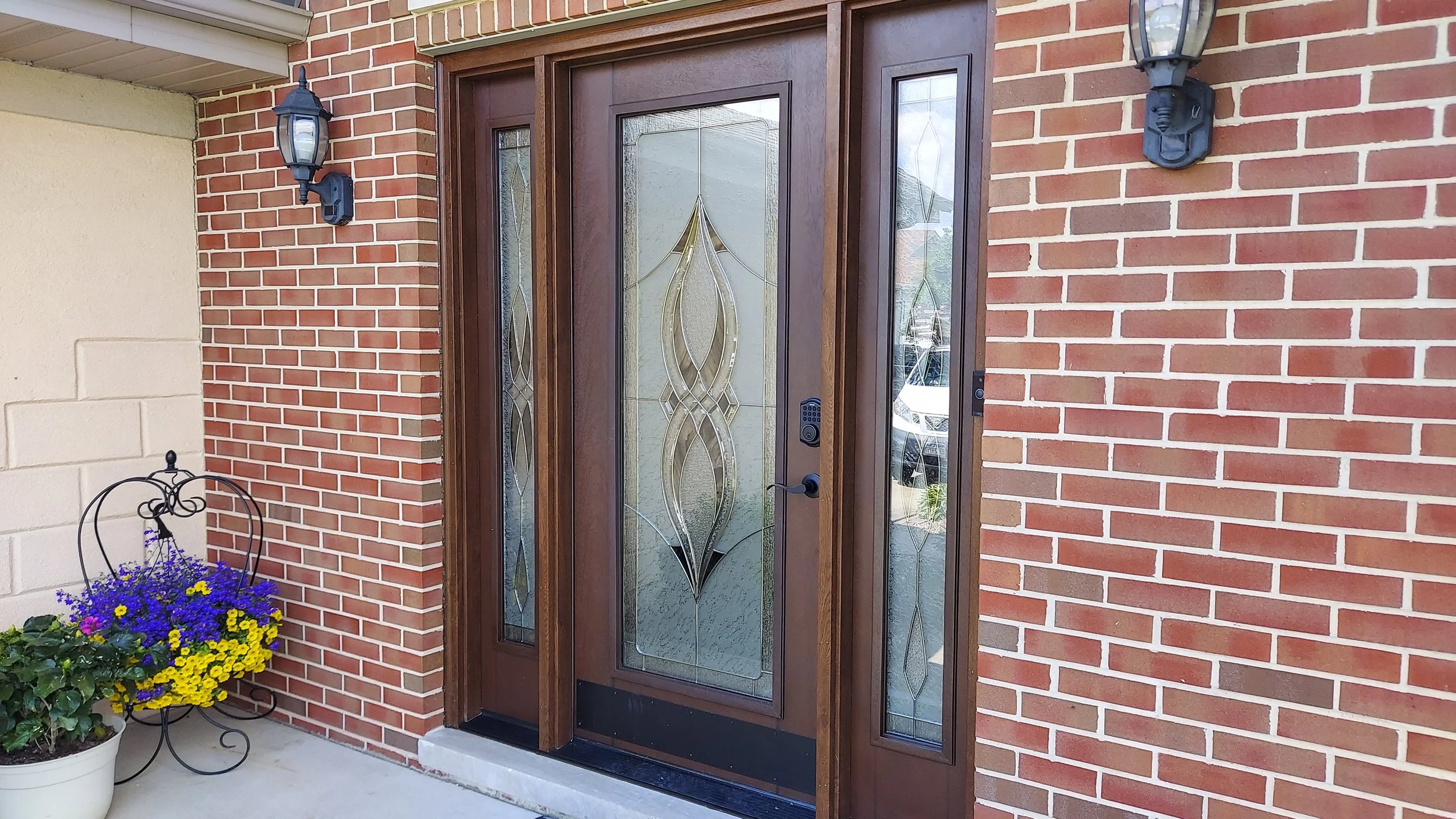 Front door with decorative glass panel, flanked by brick wall and two wall-mounted lantern lights, with a potted flower arrangement on the porch.