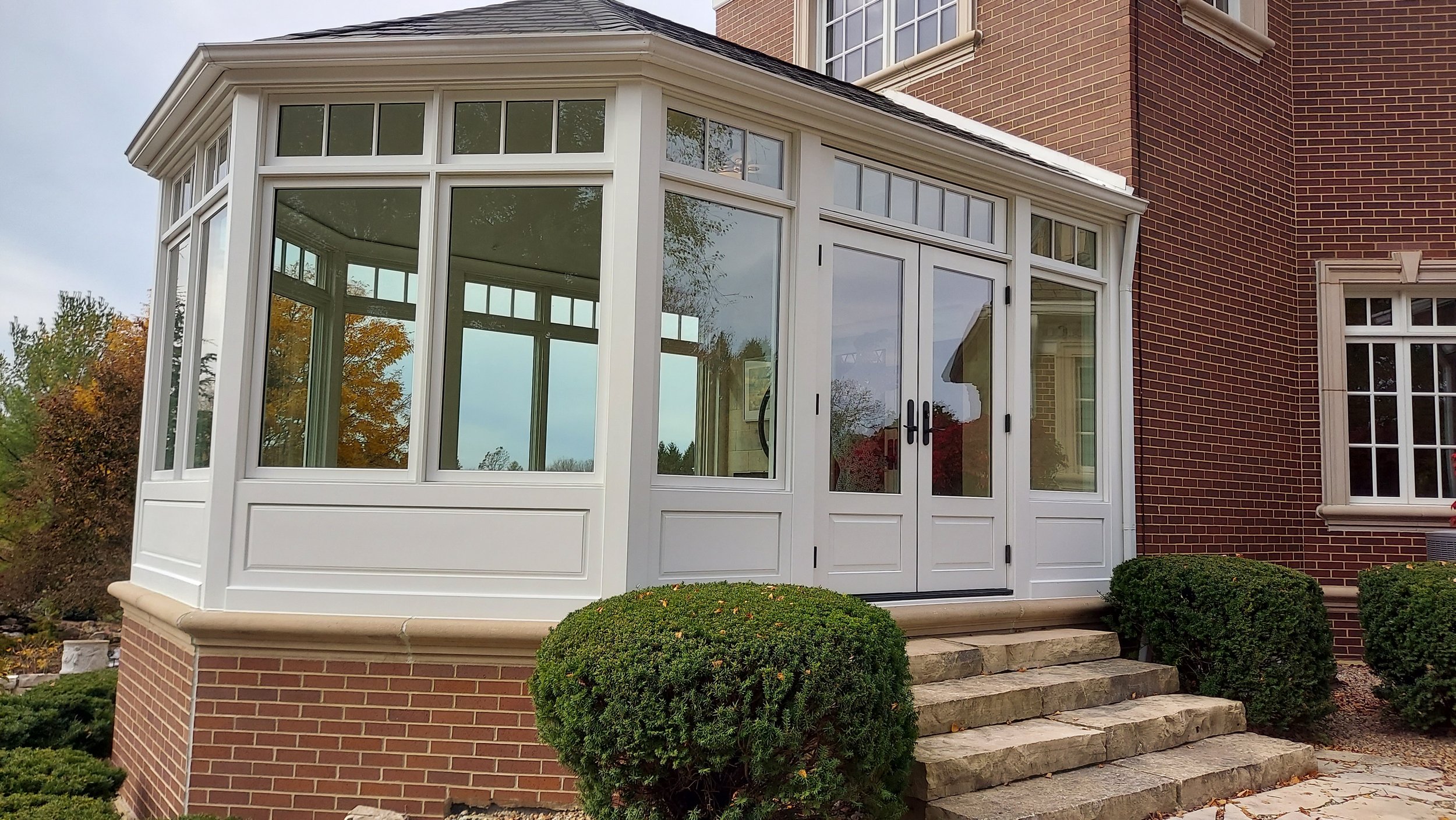 A brick house with a white enclosed porch with large windows and a glass double door, leading to stone steps and landscaped bushes.