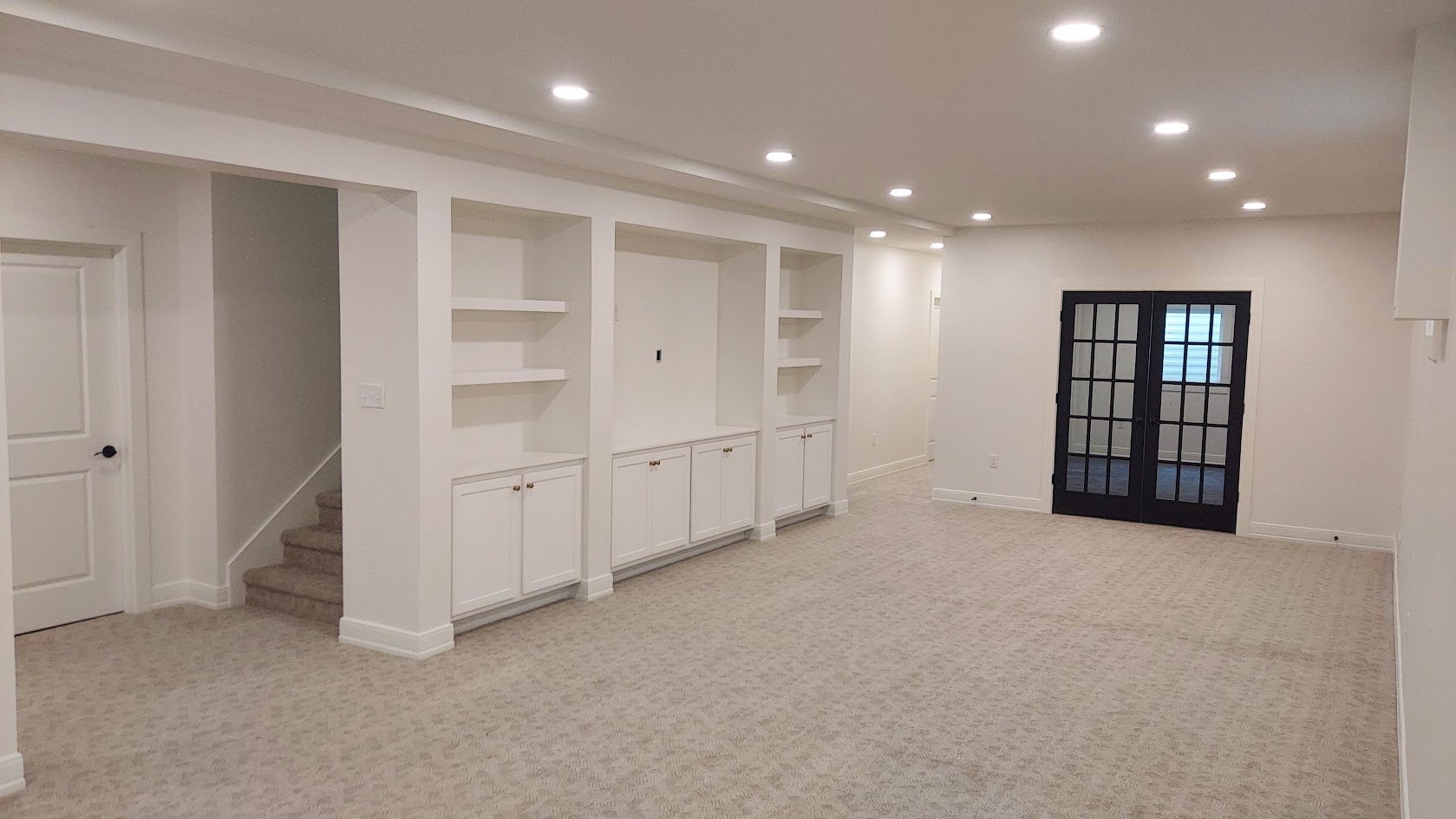 Empty living room with built-in white cabinetry, beige carpet, black glass double doors, and recessed ceiling lights.