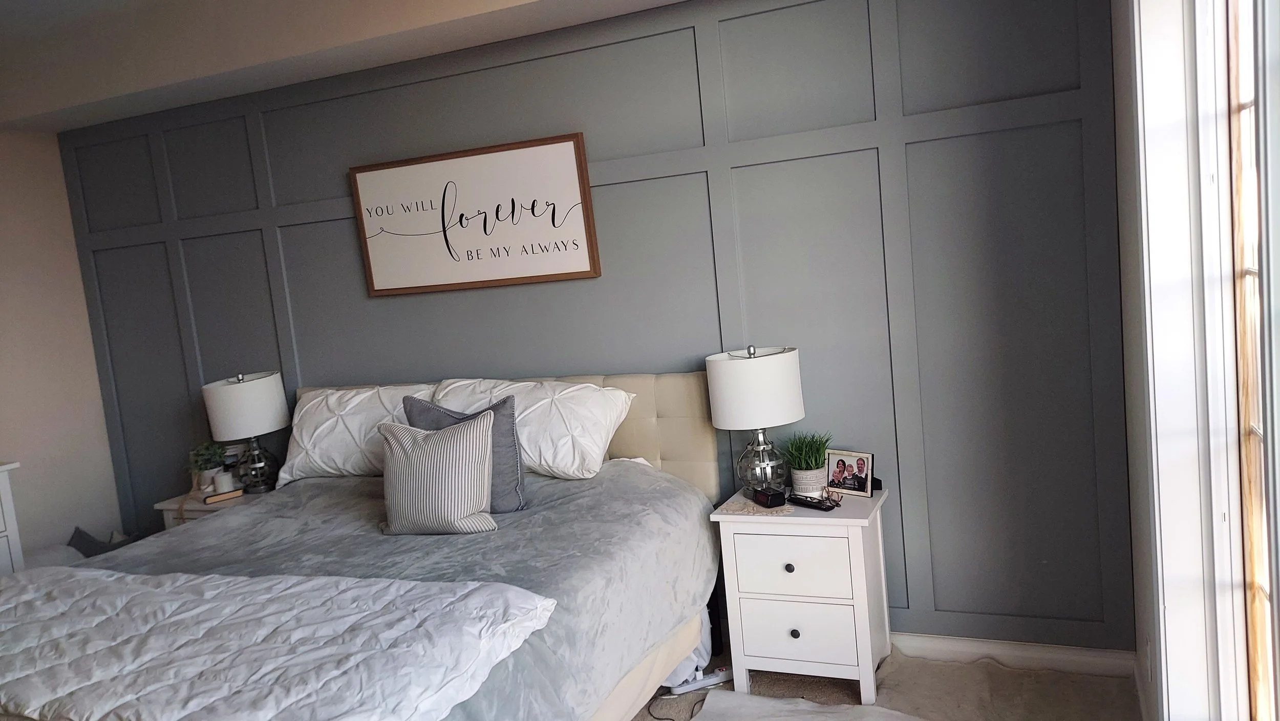 A bedroom with a bed, white nightstand, and lamps on either side of the bed. The wall behind the bed has gray paneling, and above it is a framed sign that reads, 'You will forever be my always.' The nightstand has a small plant, framed photo, and other small objects.