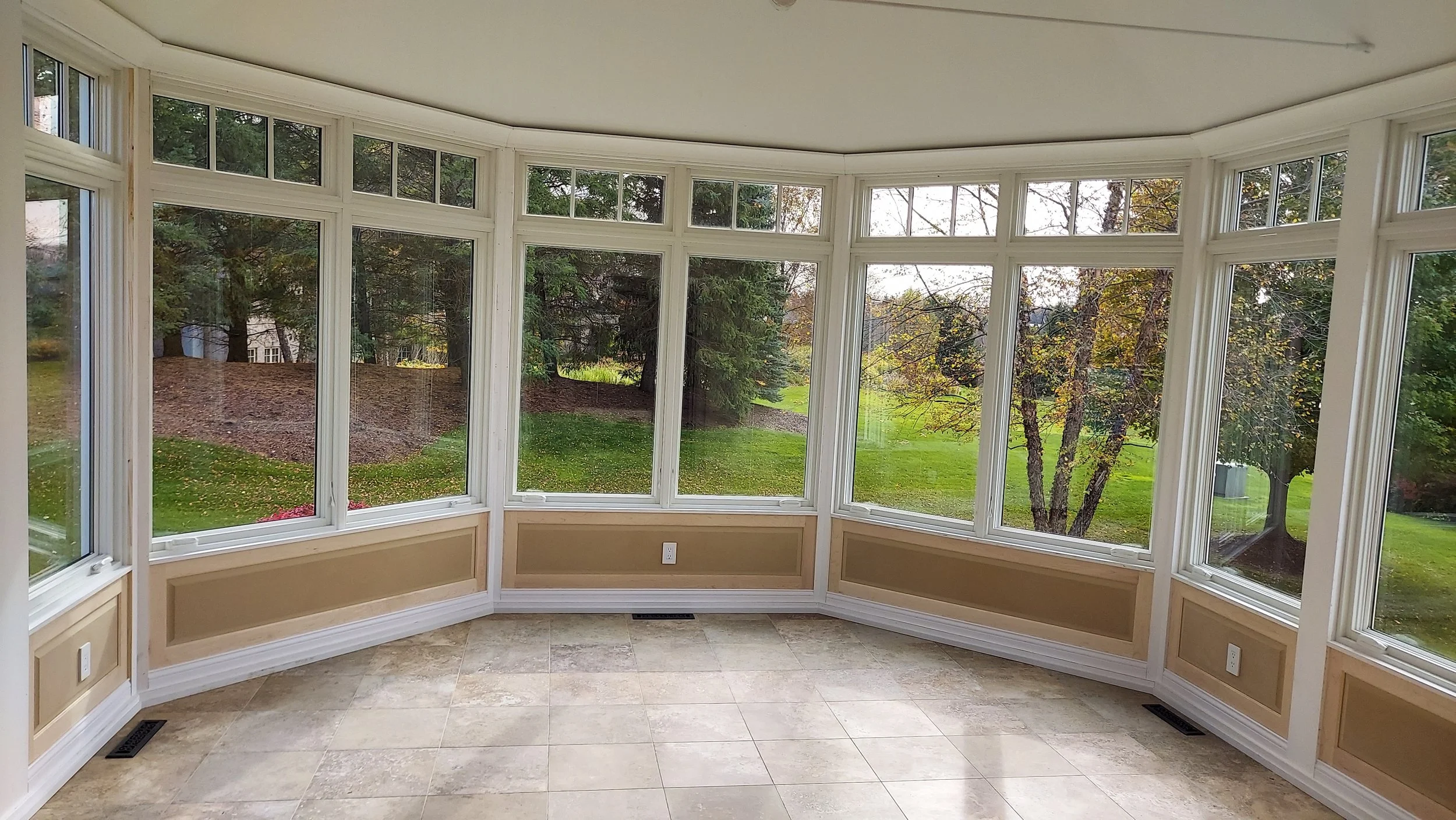 Empty sunroom with large windows overlooking a grassy yard with trees, tiled floor, and beige lower wall paneling.