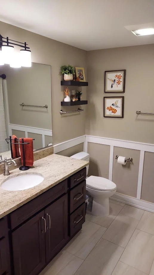 A bathroom with a granite countertop vanity, dark cabinet drawers, a large mirror, framed wall art of birds and flowers, a toilet with a toilet paper roll, and decorative shelves with plants and artwork.