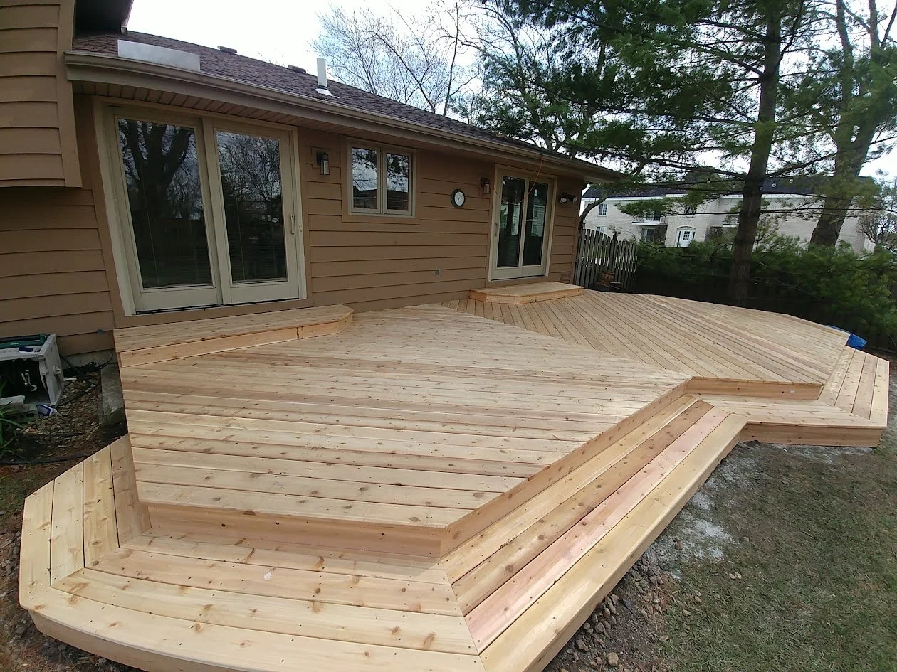 Newly built wooden deck attached to the back of a house, with stairs leading down to the yard, surrounded by a backyard with trees and neighboring houses.