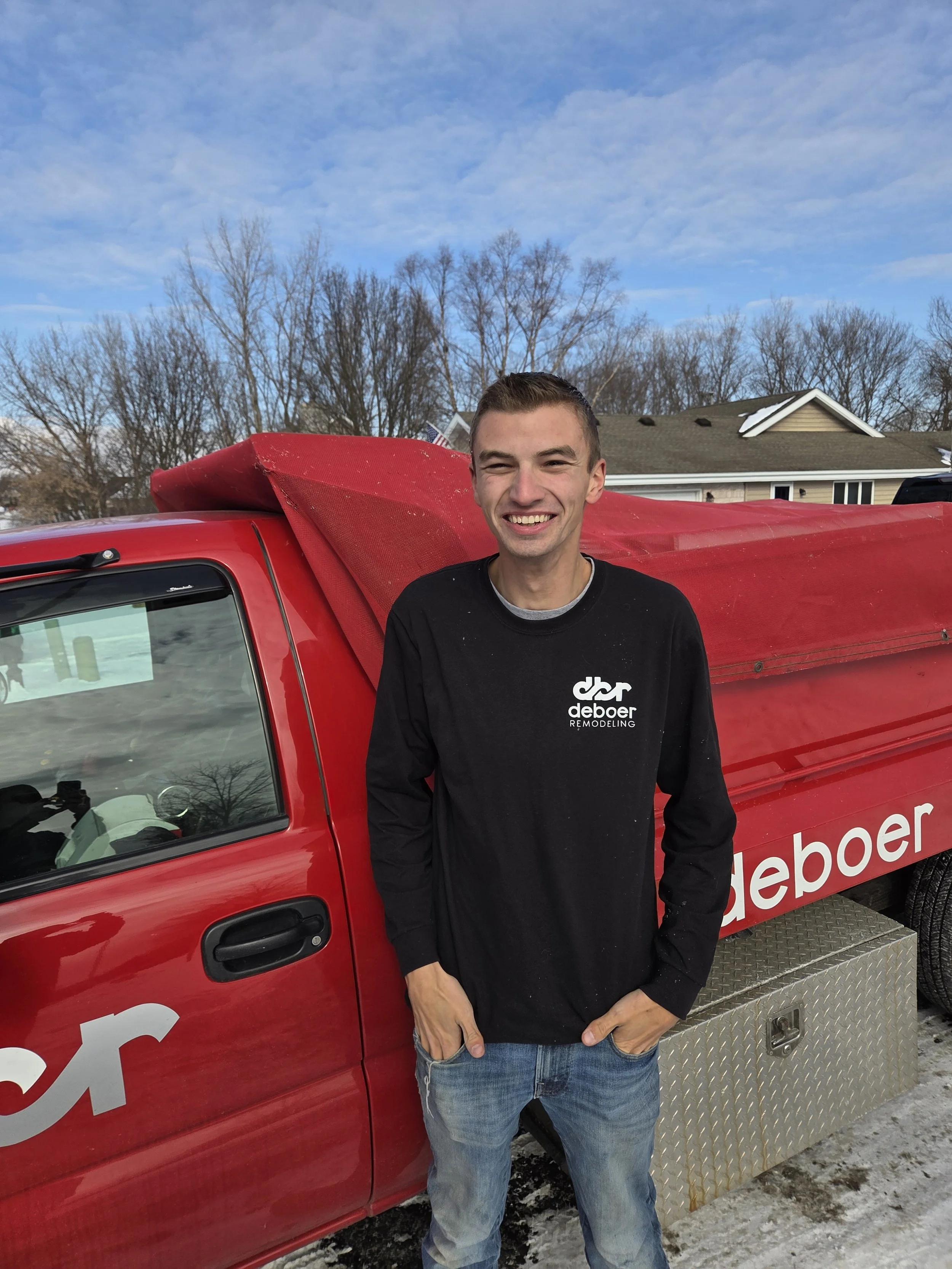 A young man standing next to a red Deboer Remodeling truck, smiling outdoors on a clear day.