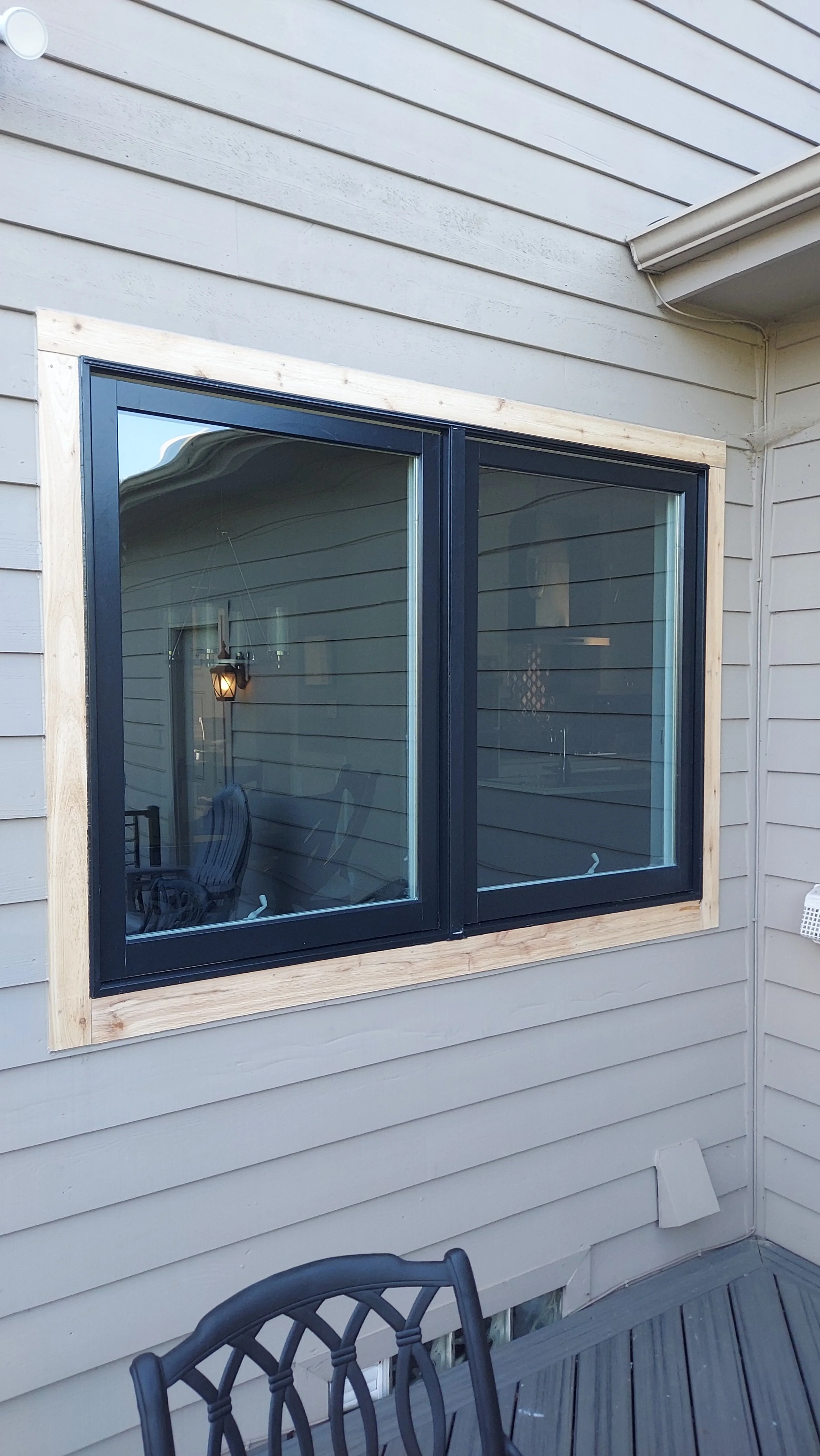 Exterior view of a house with beige horizontal siding, a black window with a wooden frame, and part of a deck with black metal chairs.