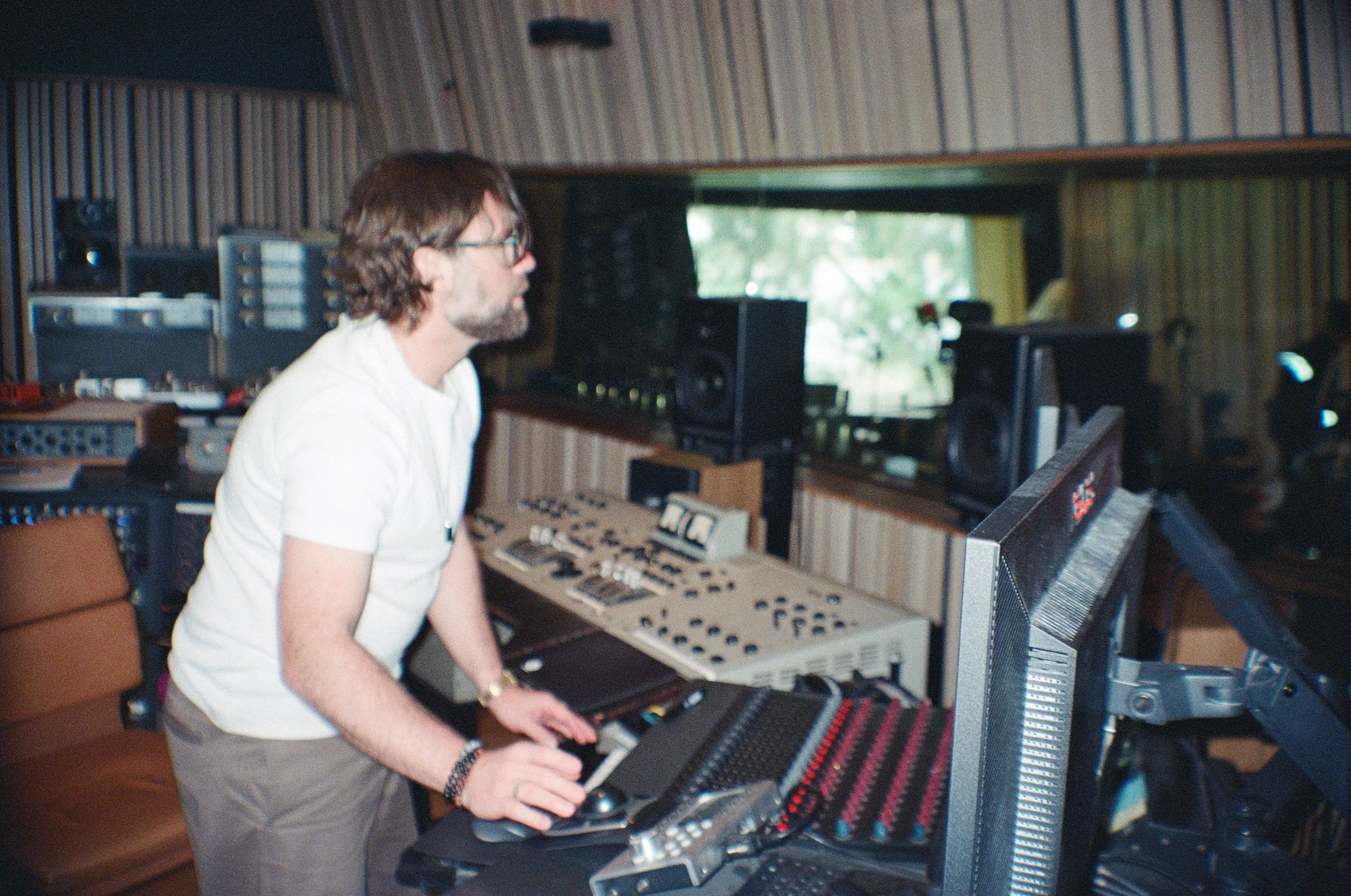 Timothy McArtney with glasses and a beard wearing a white shirt working in a recording studio surrounded by audio equipment, including a keyboard, speakers, and mixing consoles.