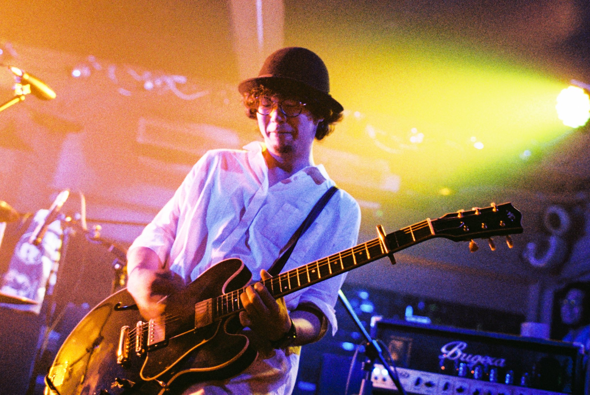 Person playing electric guitar on stage wearing a hat and glasses, under colorful stage lights.