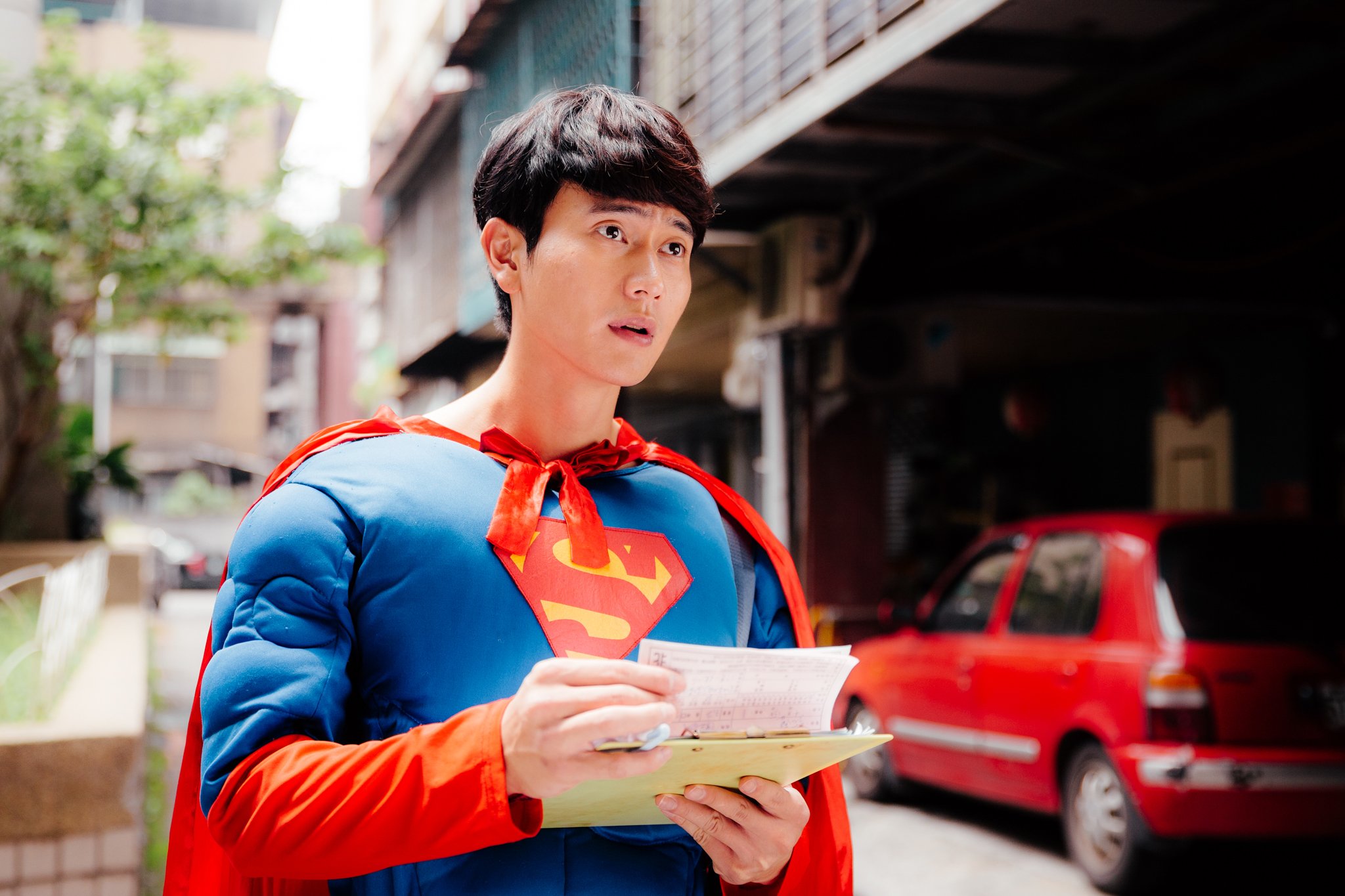 Person wearing Superman costume holding a clipboard on a street with a red car in the background.