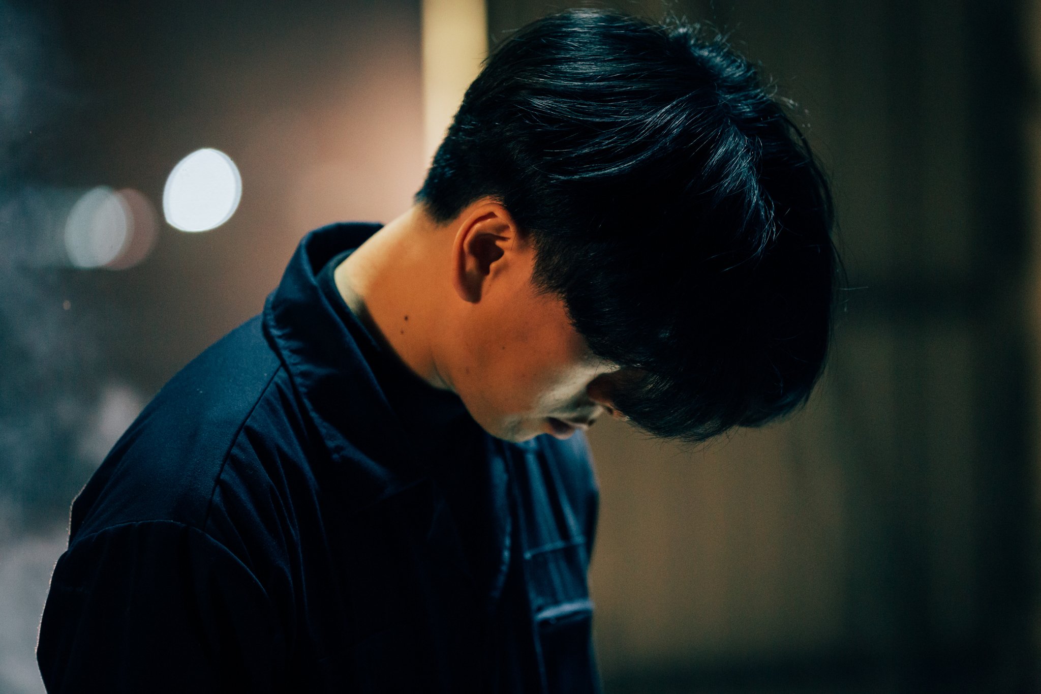 Person with dark hair looking down, wearing dark clothing, in dim lighting.