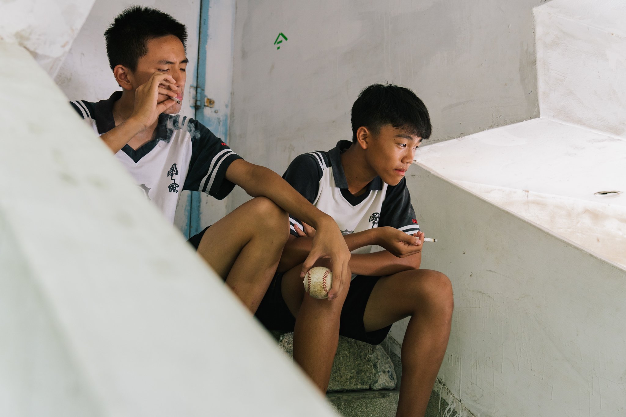 Two young boys, wearing sports uniforms, sitting on stairs, one holding a baseball, both smoking cigarettes.