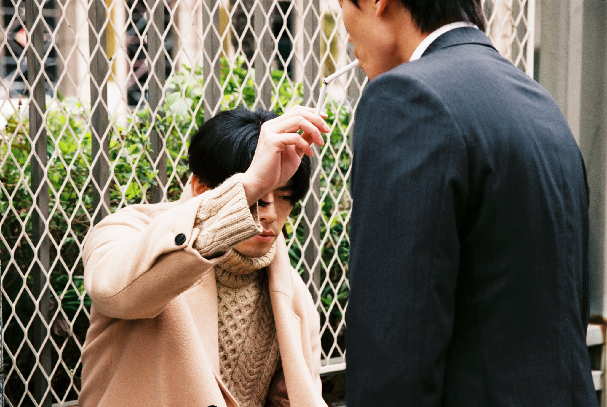 Two men interacting outdoors near a metal fence; one man in a beige coat and sweater is lighting a cigarette for a man in a suit.