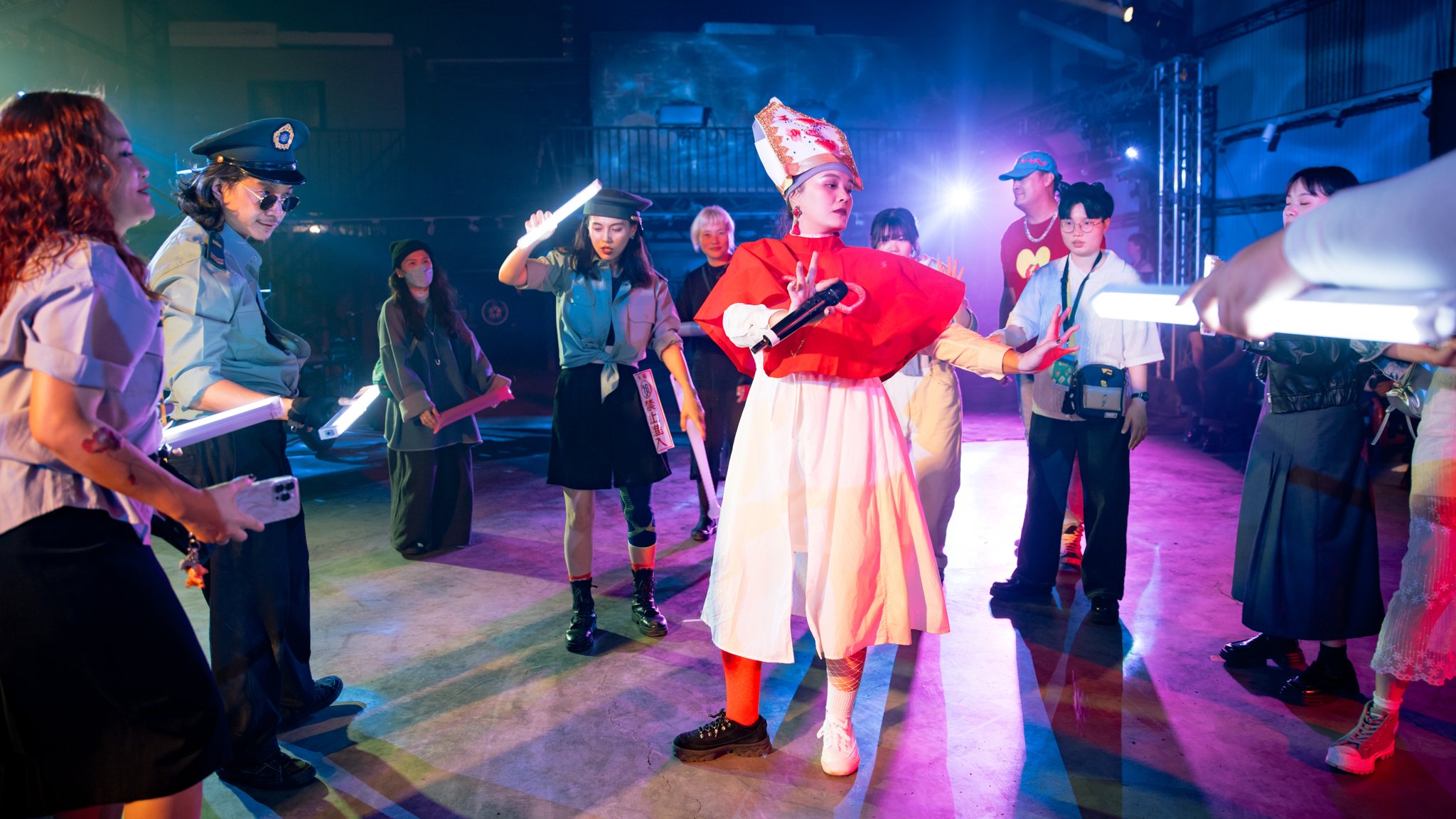 A group of people in theatrical costumes and uniforms, with one person in the center wearing a red and white outfit and holding a microphone, surrounded by others holding white light sticks, in a dimly lit setting with colorful lighting.
