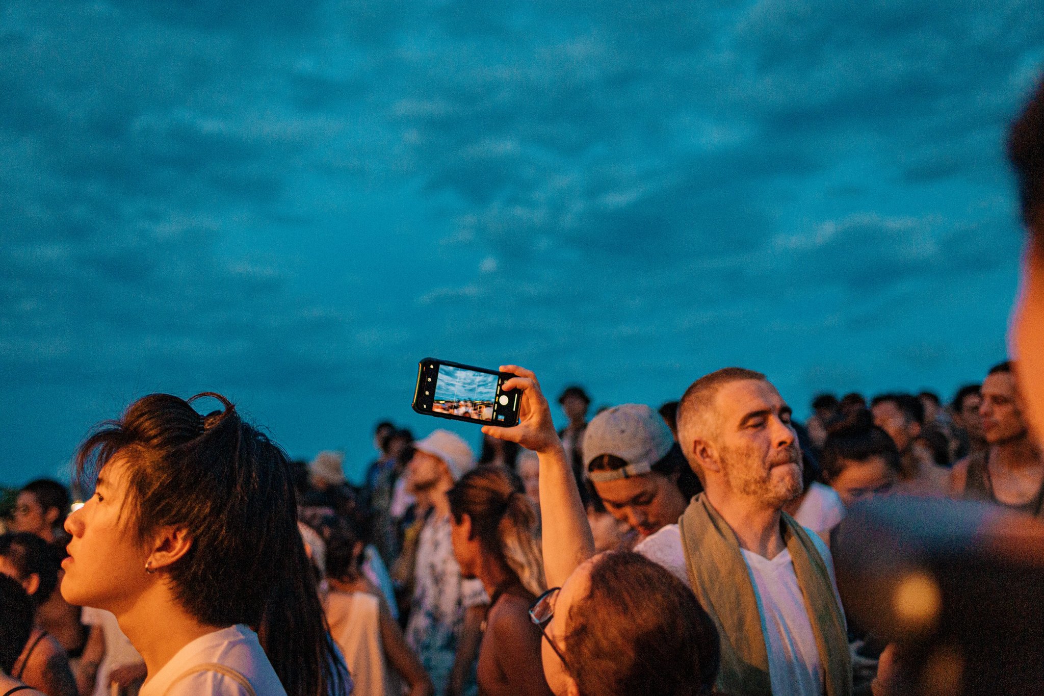 A crowd enjoying a festival or outdoor event, with a person holding up a smartphone to take a photo, beneath a blue sky.