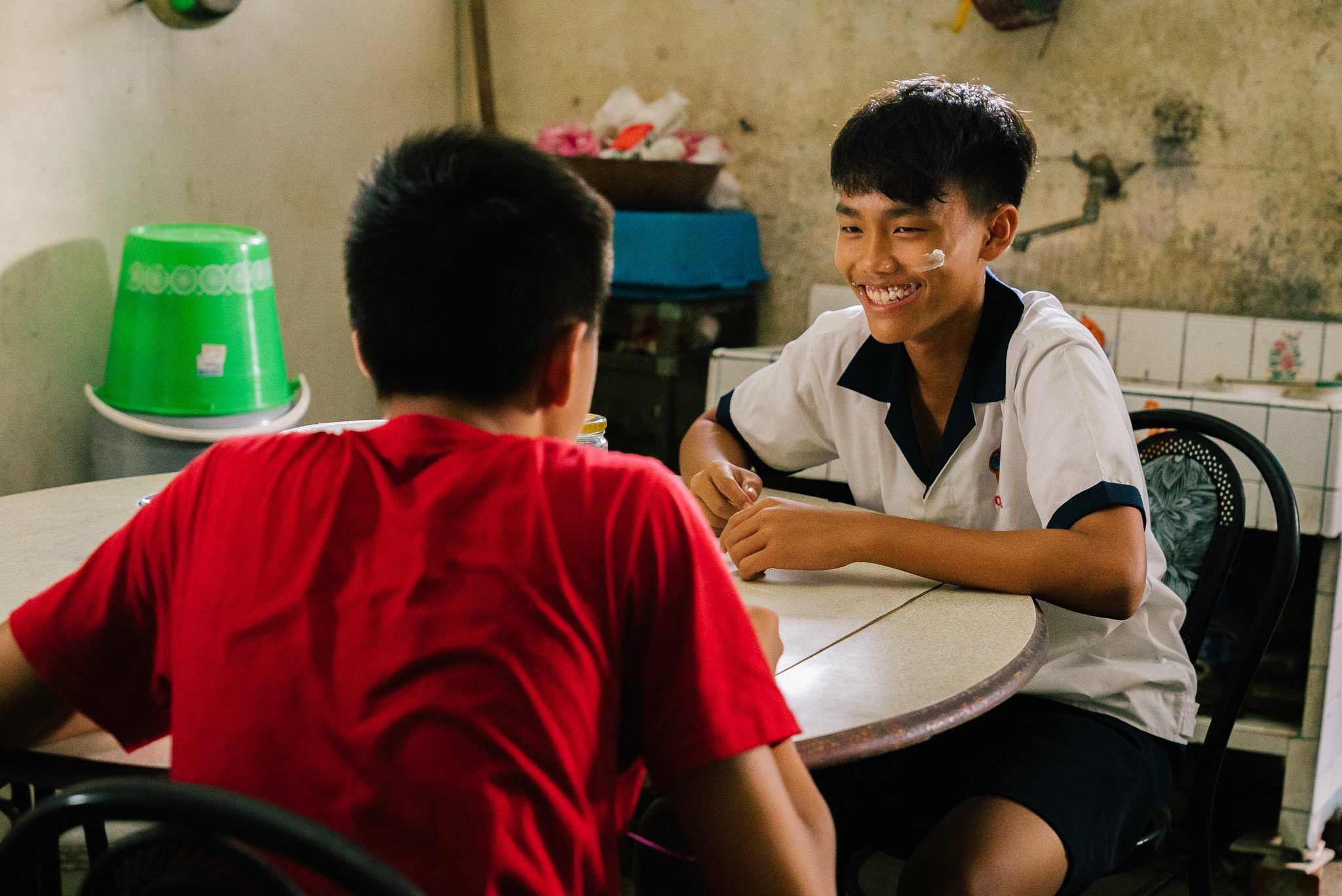 Two young boys sitting at a table indoors, one wearing a red shirt and the other in a white shirt with dark trim, smiling and talking.