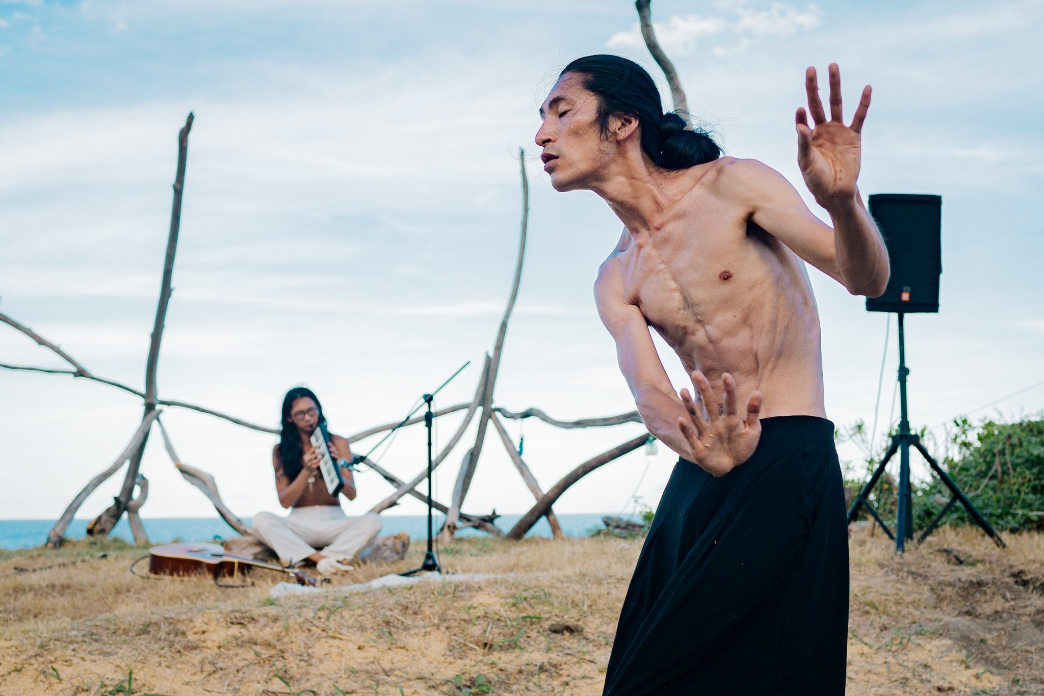 Man dancing shirtless outdoors, another person playing a melodica in background, surrounded by natural wooden structures and sound equipment.
