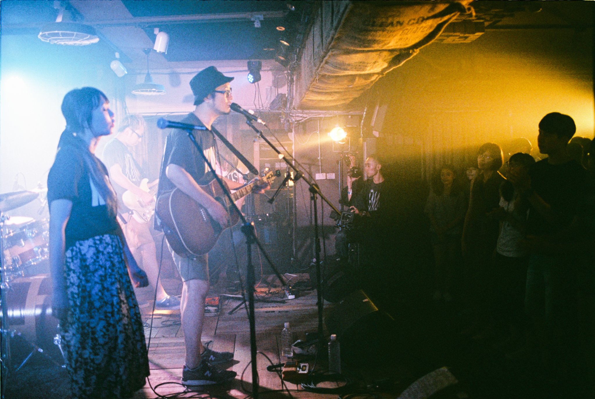 Indoor live music performance featuring a band on stage with a guitarist and vocalist. Audience watching under atmospheric lighting and haze.