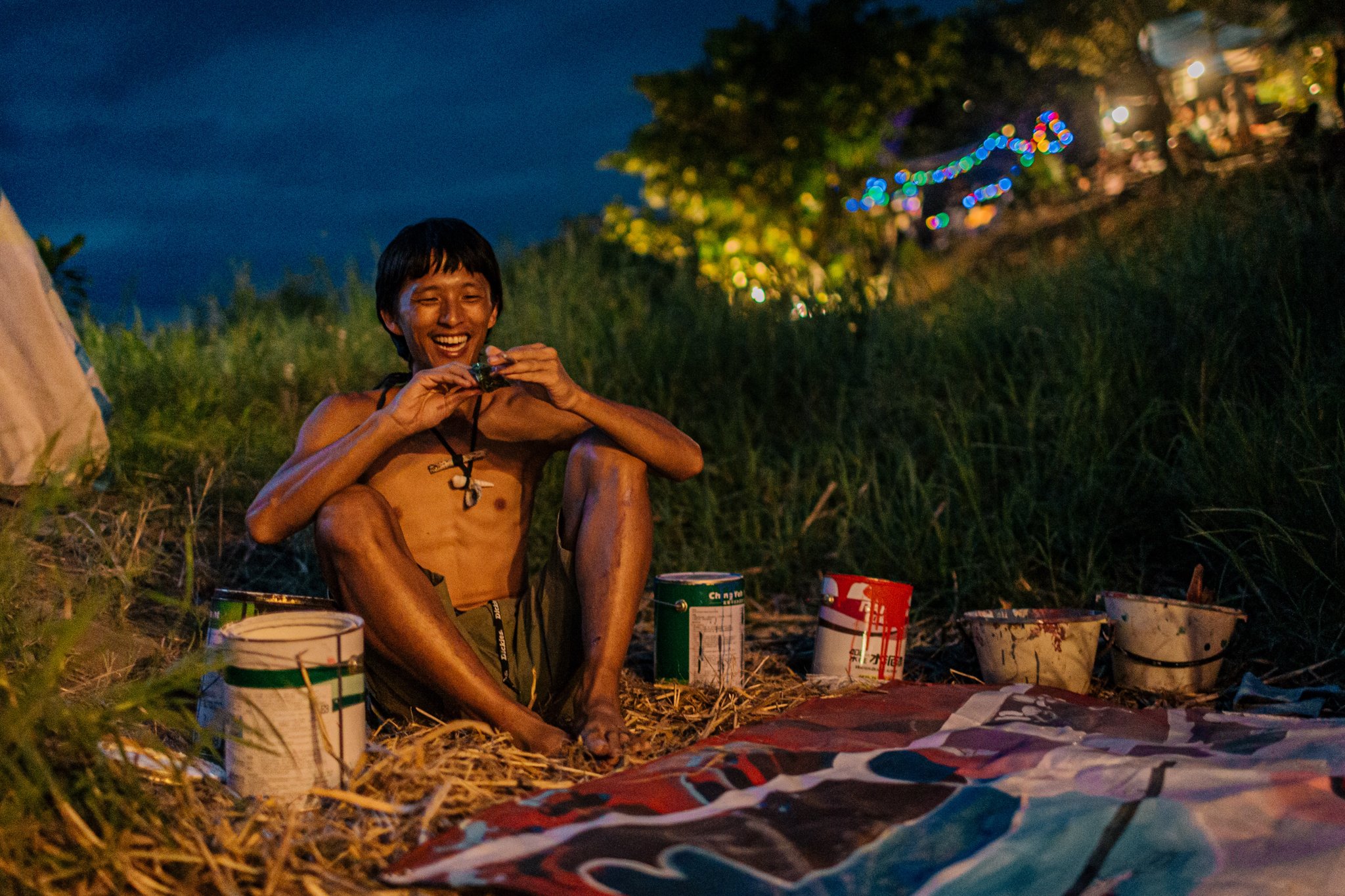 Man sitting outdoors at night, surrounded by paint cans, holding an instrument, with colorful lights in the background.