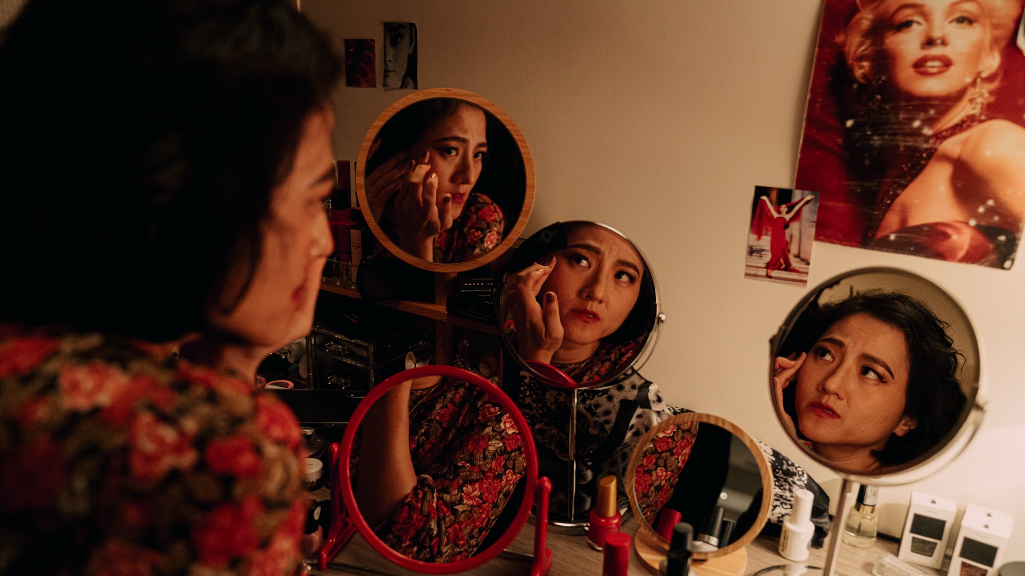 Woman applying makeup in front of multiple mirrors on a vanity table, decorated with posters and cosmetics.
