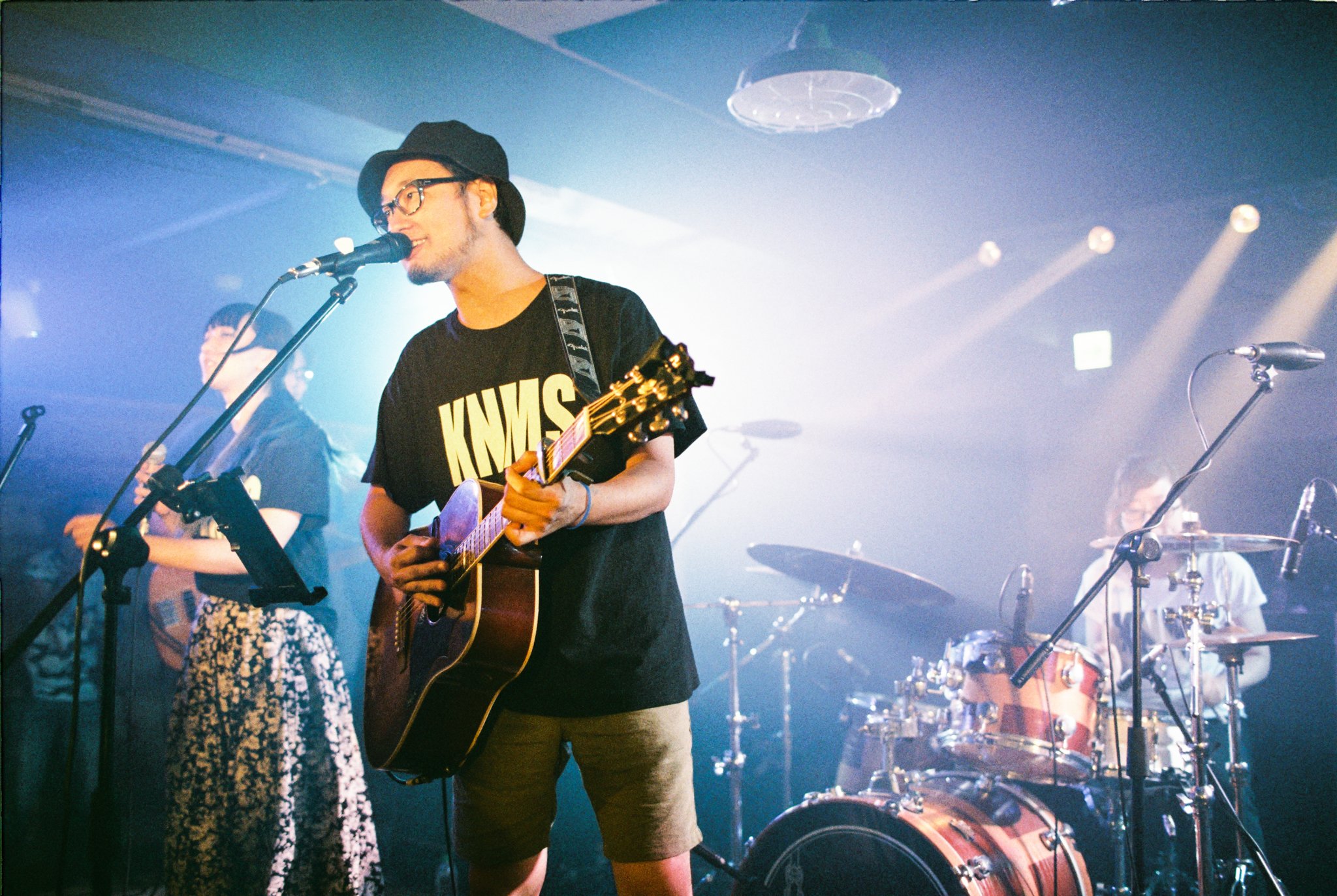 A band performing live on stage, featuring a guitarist and singer wearing a "KNMS" shirt, with other band members playing instruments in a dimly lit venue.