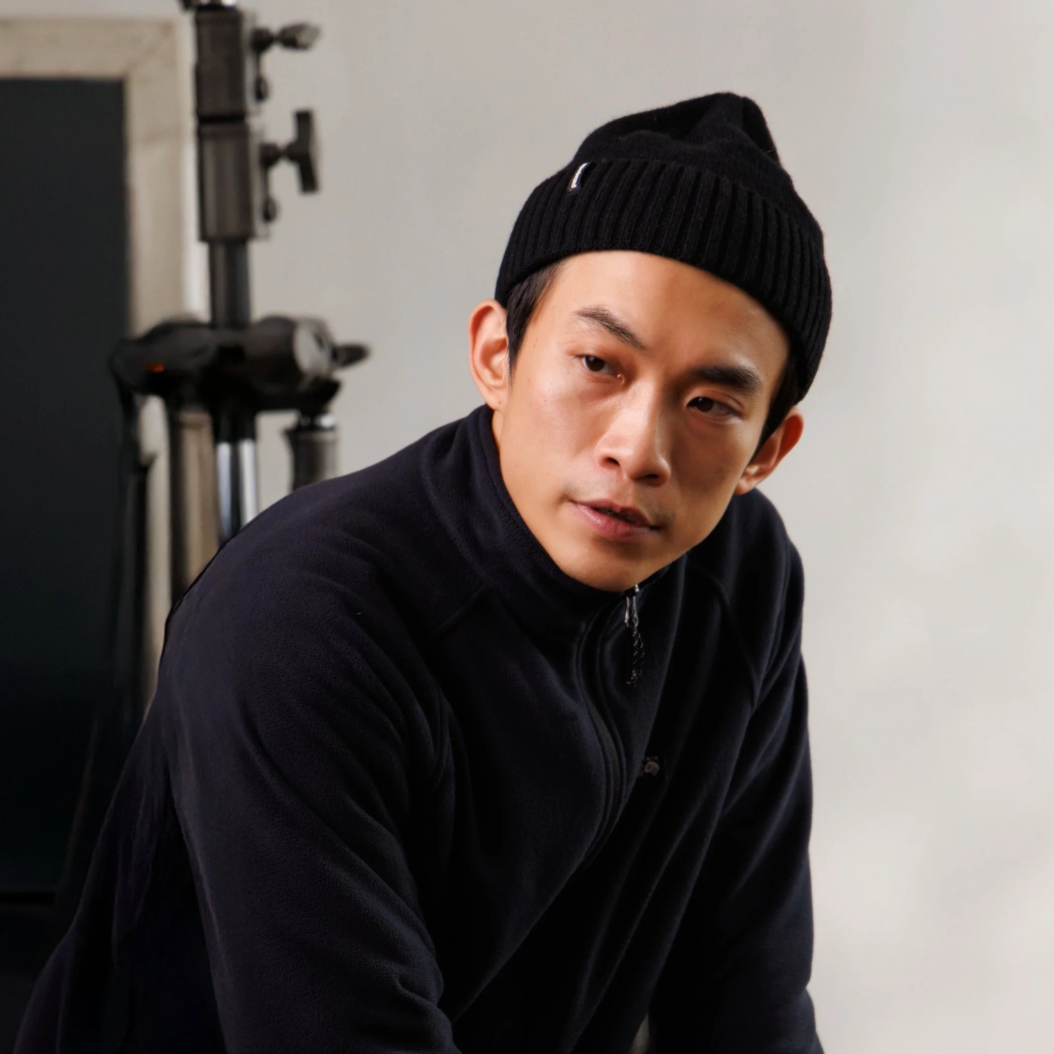 Man wearing a black beanie and dark jacket indoors, with photographic equipment in the background.