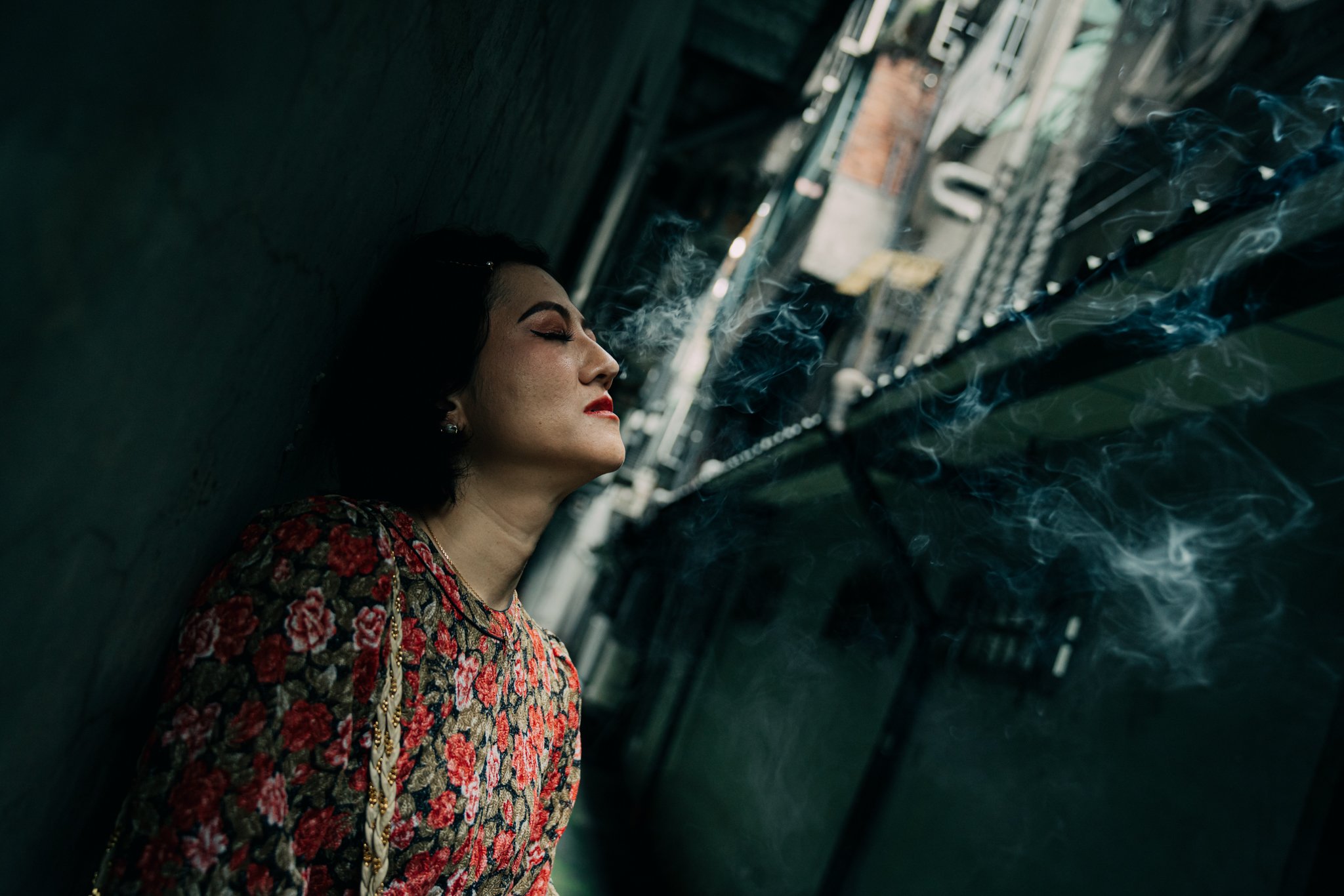 Person in floral dress leaning against a wall with eyes closed and smoke around.