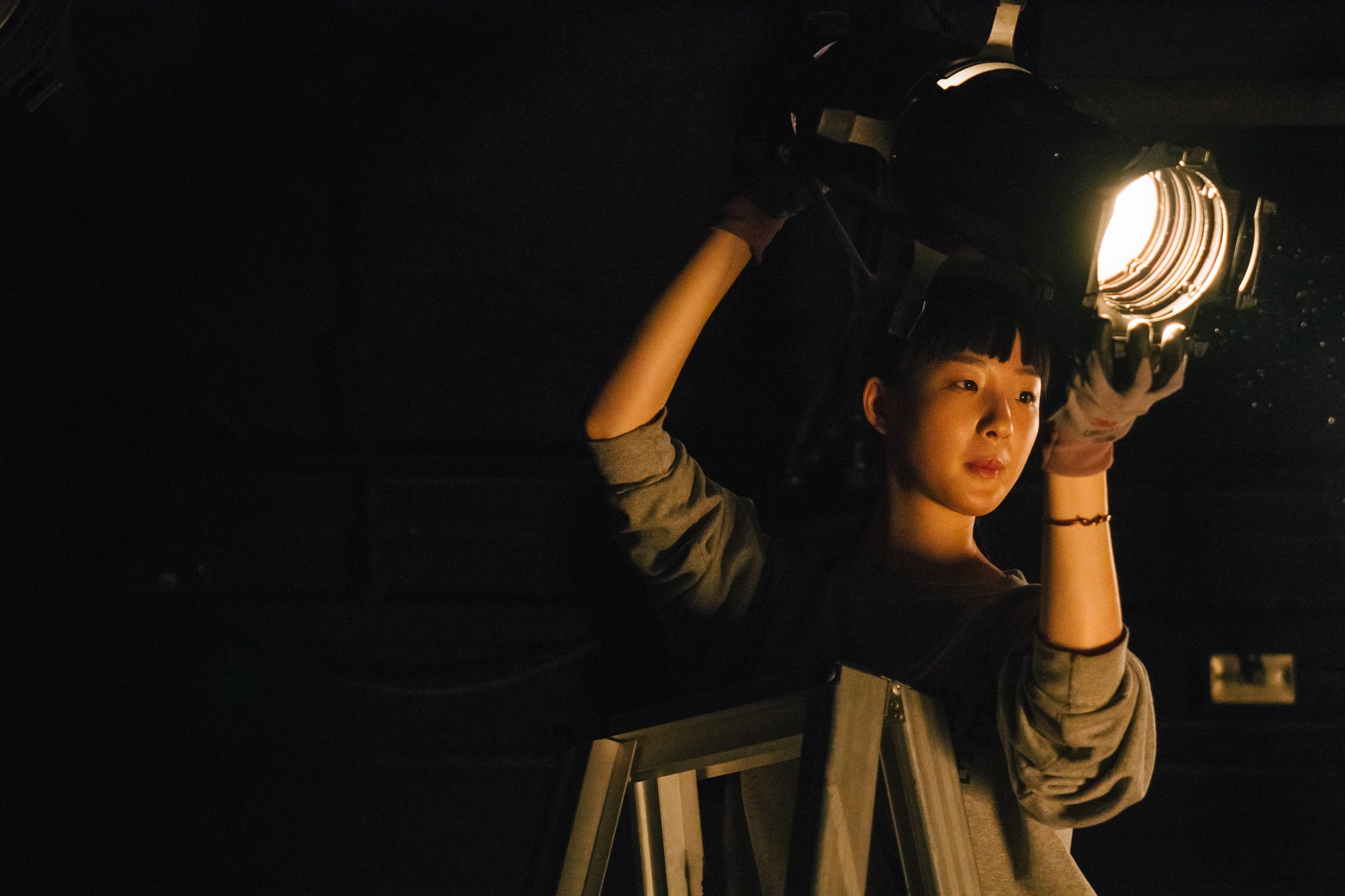 Person adjusting stage light in a dark room, standing on a ladder.