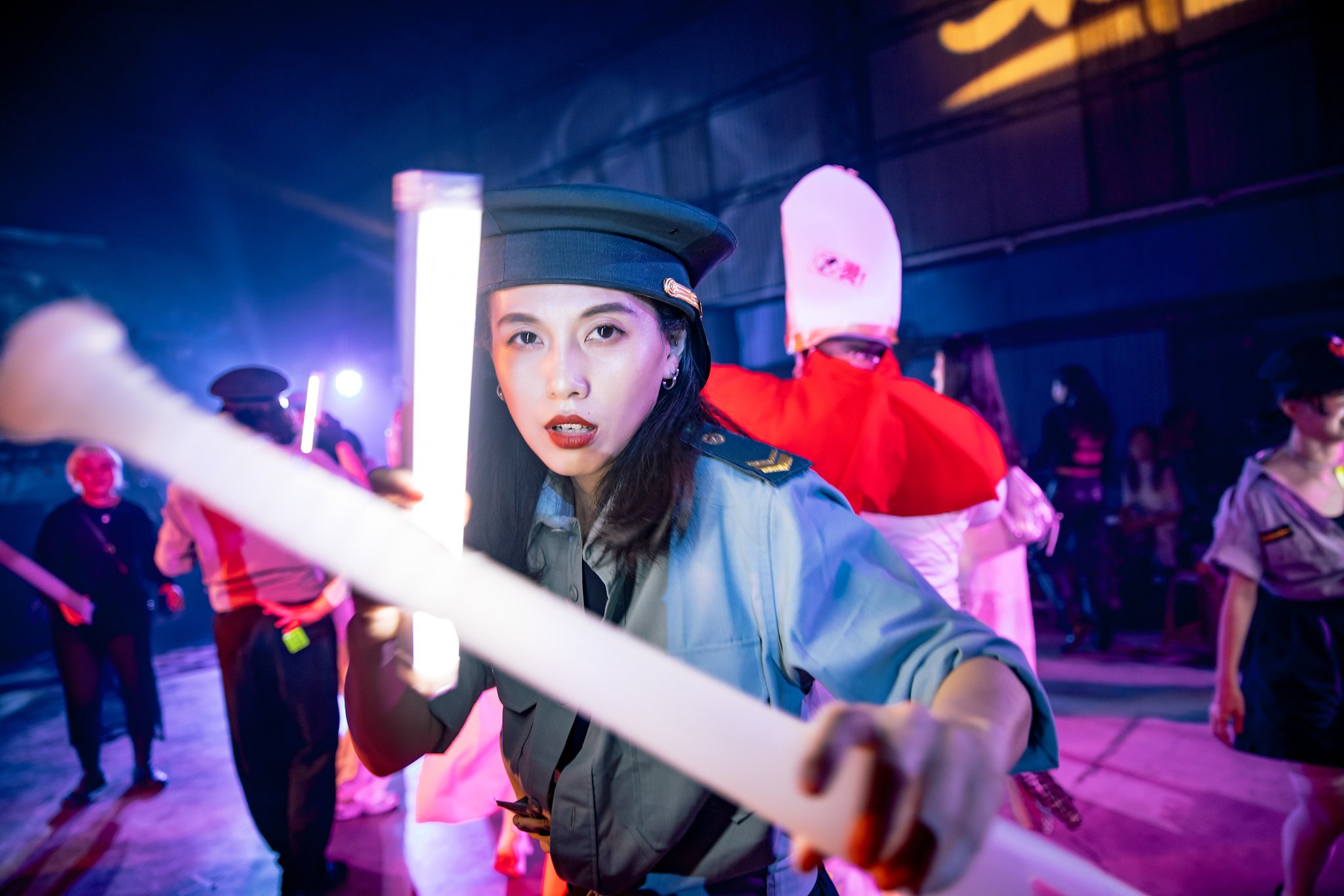 People in colorful costumes and uniforms holding illuminated batons, in a dimly lit indoor setting.
