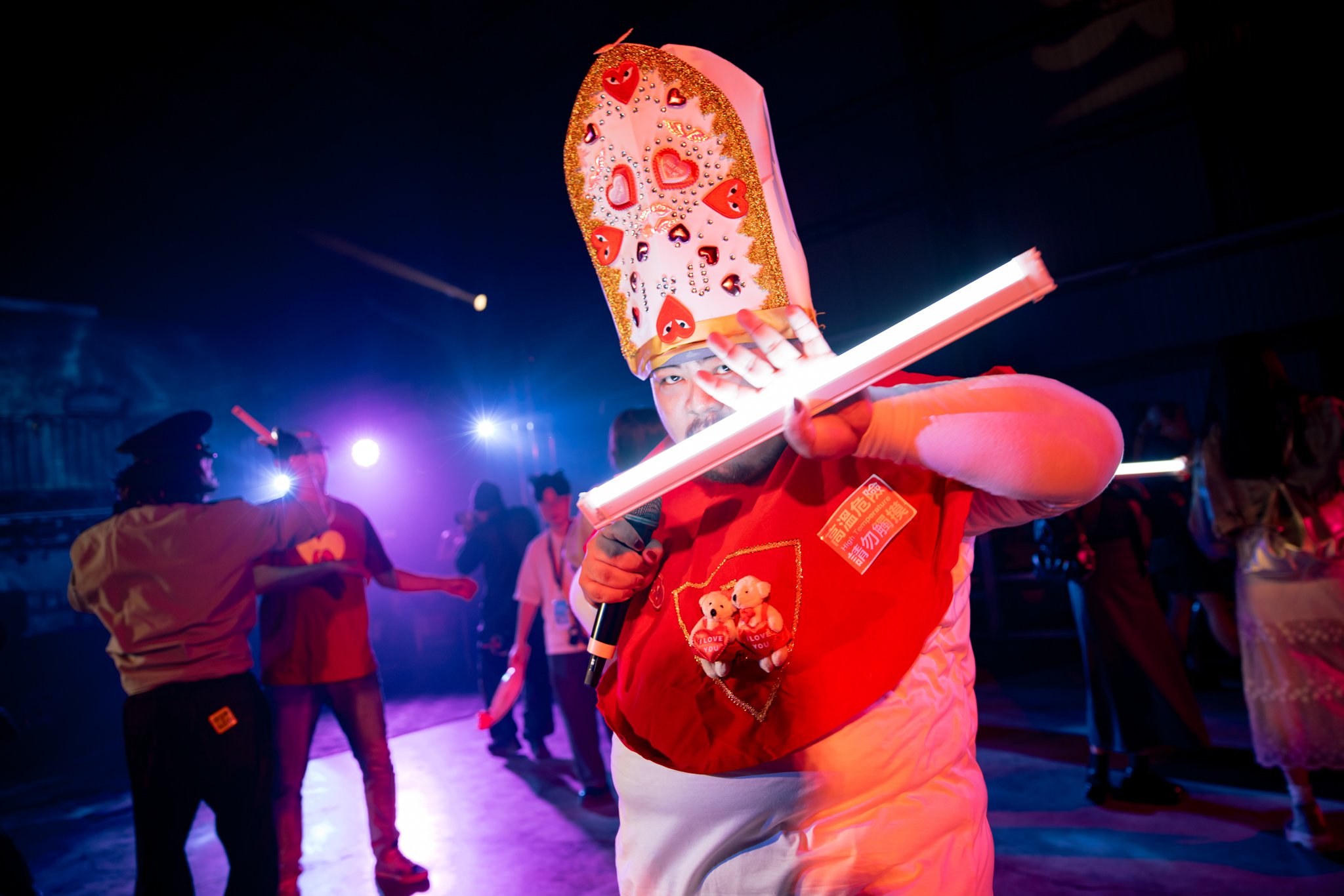 Person in a colorful costume with heart decorations, holding a glowing stick, in a lively event with colorful lights and people dancing in the background.