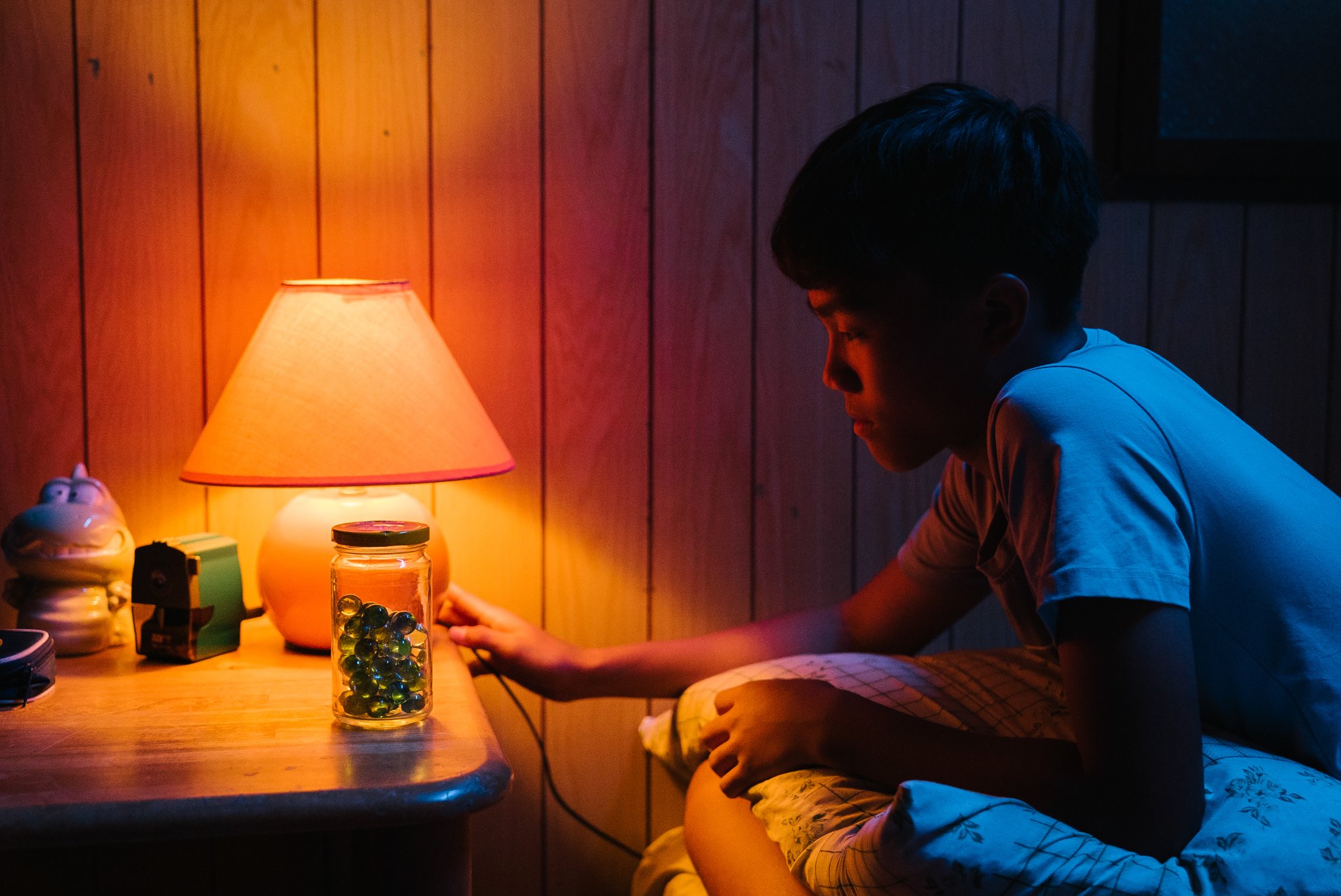 A person sitting in a dimly lit room next to a wooden table with a lamp, a jar of marbles, and other items on it.