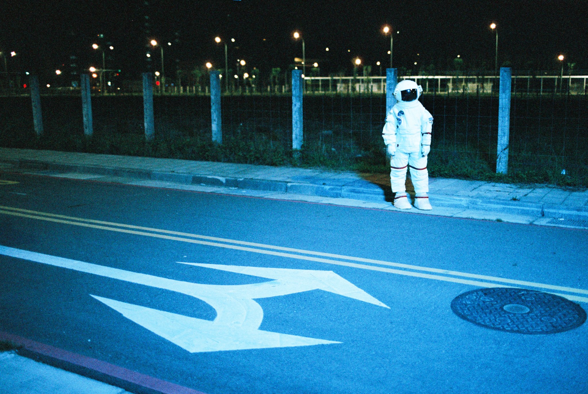 Astronaut in spacesuit standing on a city street at night, with road markings and streetlights visible.