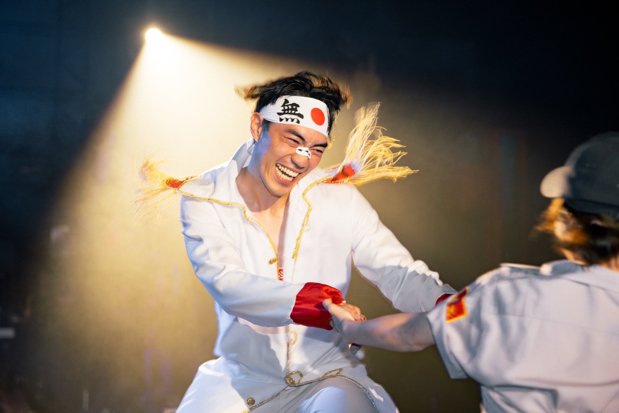 Man in costume with headband laughing and holding hands with another person on stage.