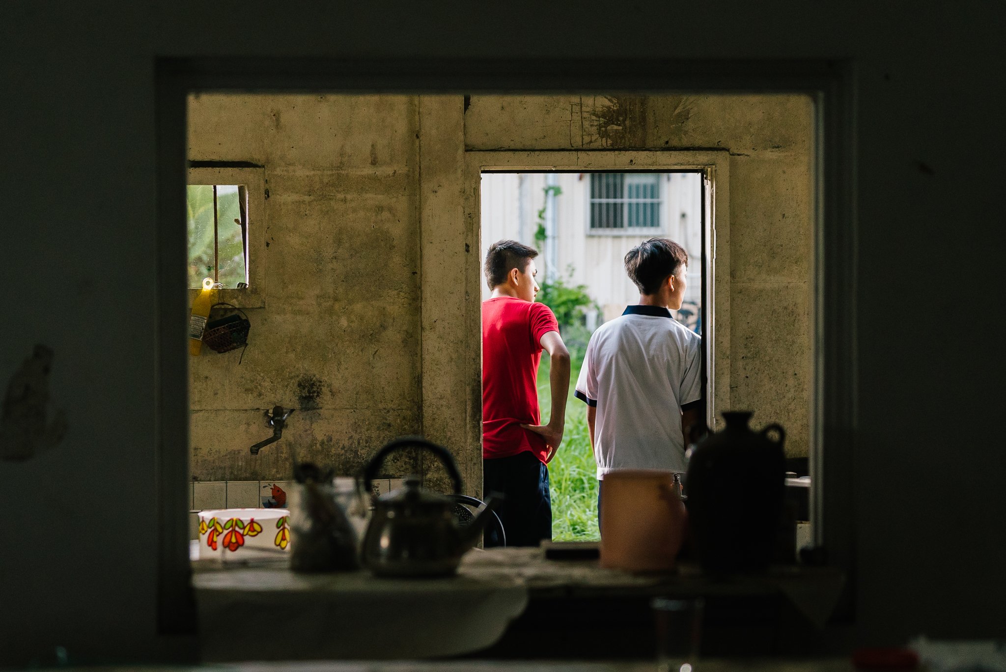 Two people standing at an open doorway, viewed from a dimly lit room with a kitchen counter in the foreground, and looking outside towards a garden or yard area.