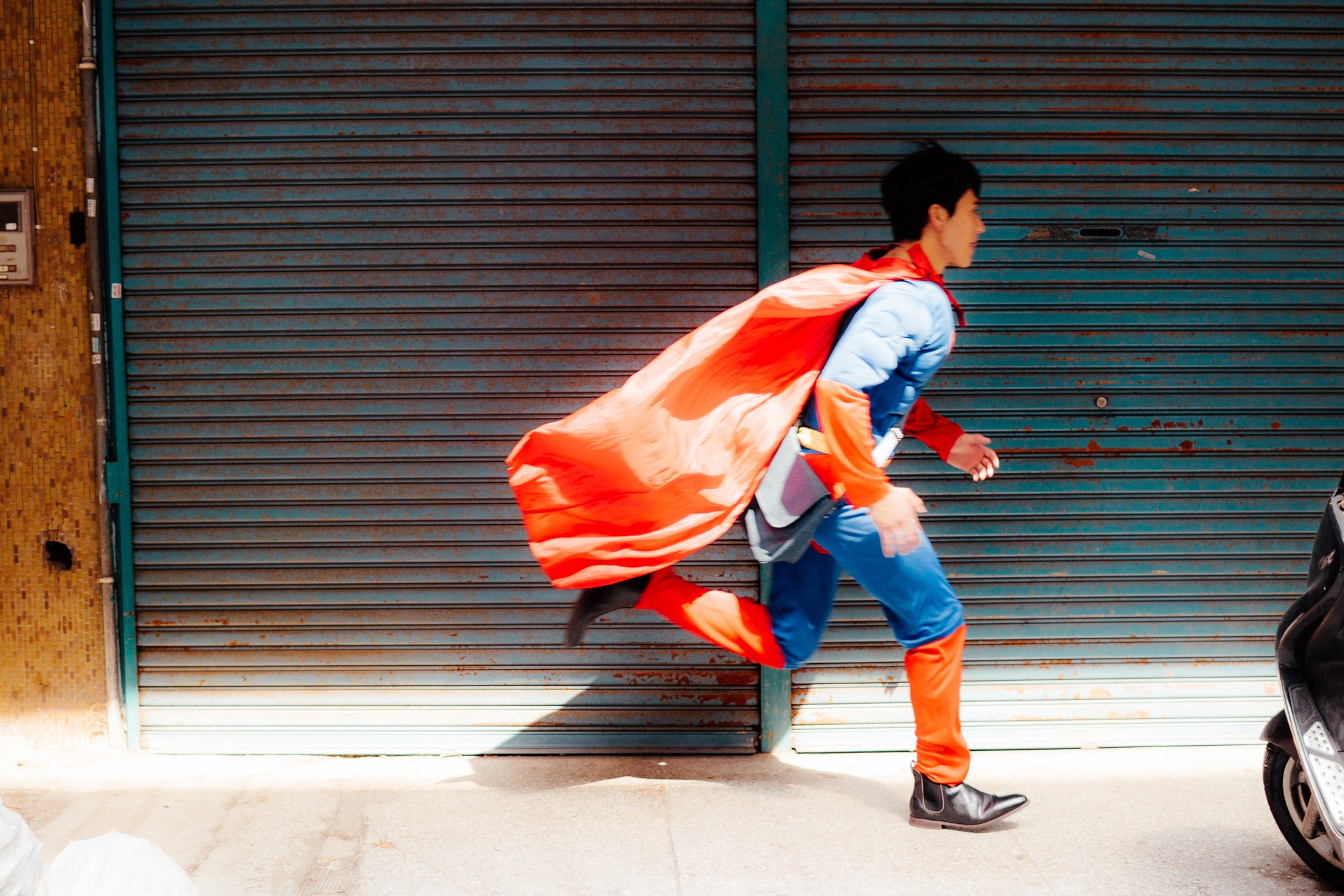 Person in superhero costume with red cape running past a closed shutter in an urban setting.