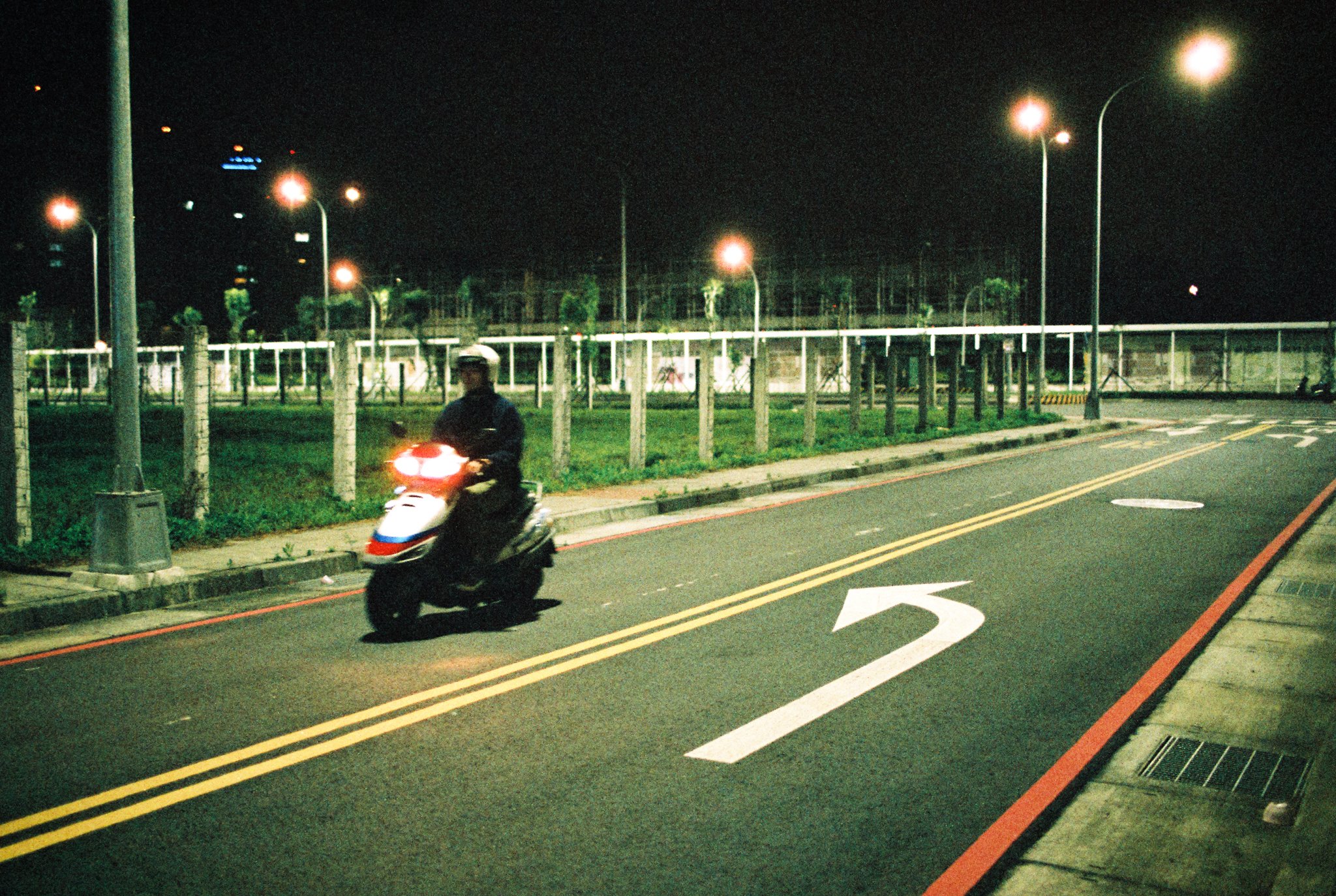 A person riding a scooter on a dimly lit street at night with light poles and a bending arrow painted on the road.