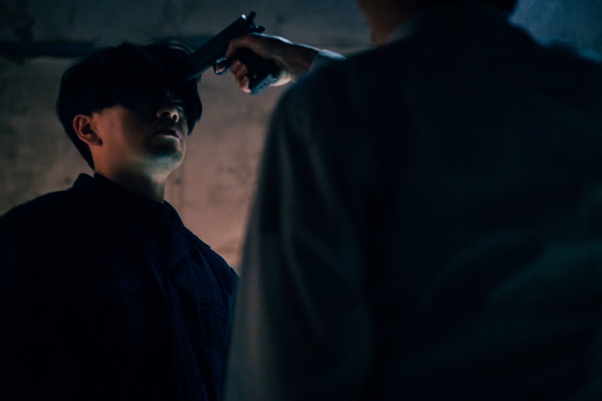Two people in a dimly lit room, one holding a gun to the other's forehead.