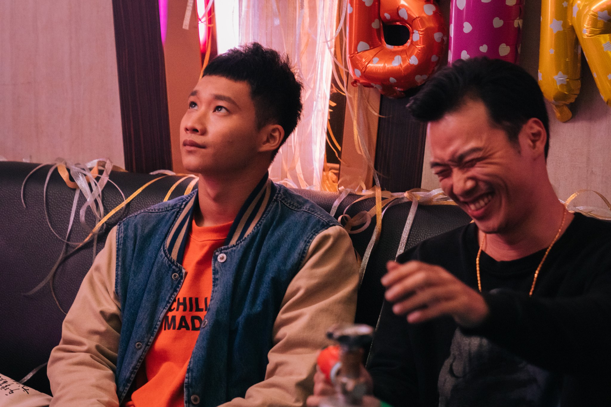 Two people sitting on a couch at a party, one looking up thoughtfully and the other laughing, with balloons and streamers in the background.