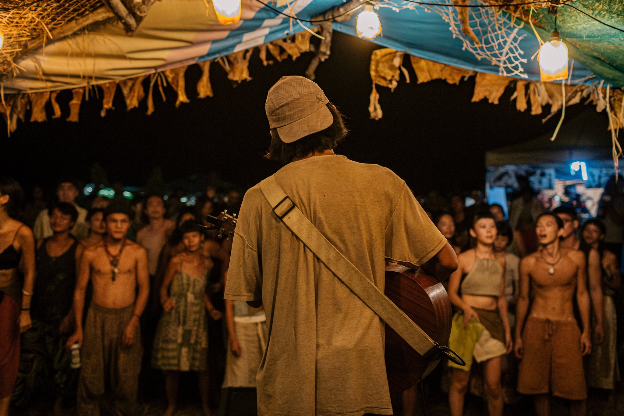 Musician playing guitar, performing to a crowd under a tent with hanging lights and decorations at night.