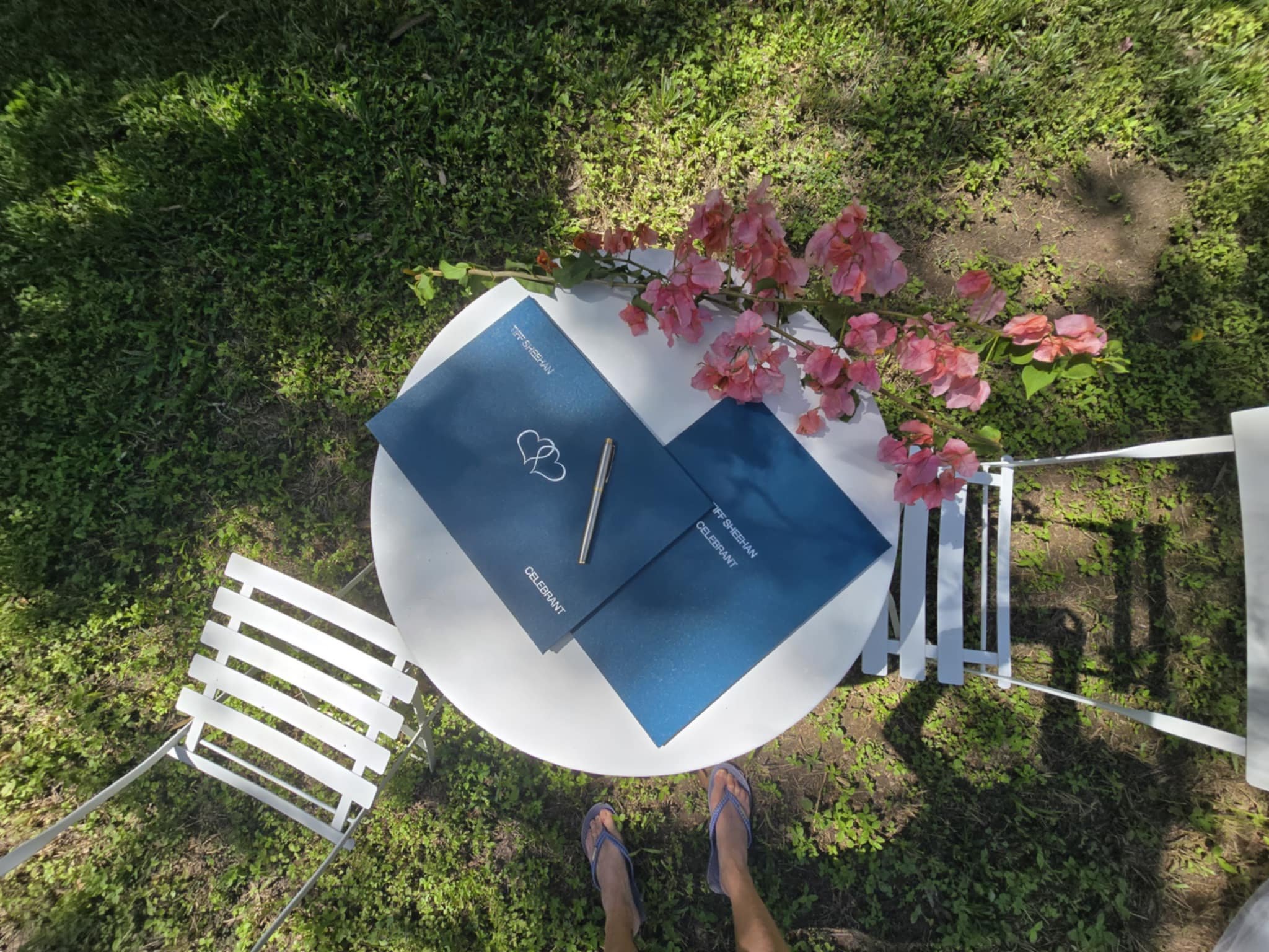 Top-down view of a small white round table with a pink flower branch, two blue cards, a gold pen, and two white chairs on grass.