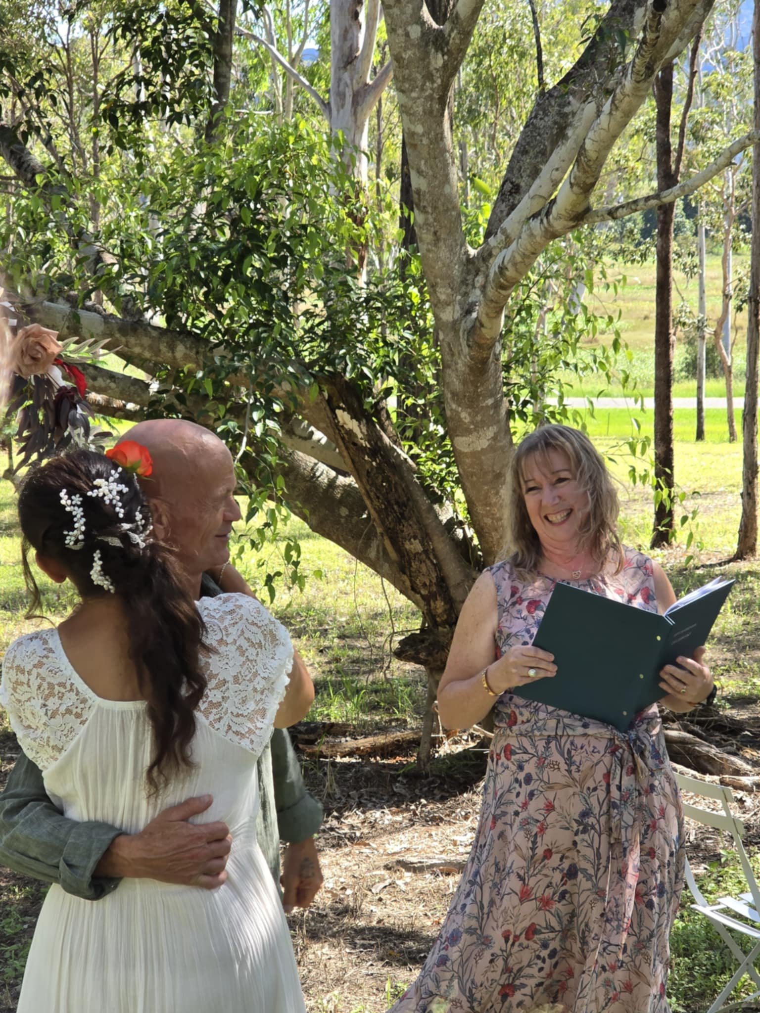 A woman reading from a book outdoors, smiling, with a man and a woman beside her under a tree in a green, sunny area.