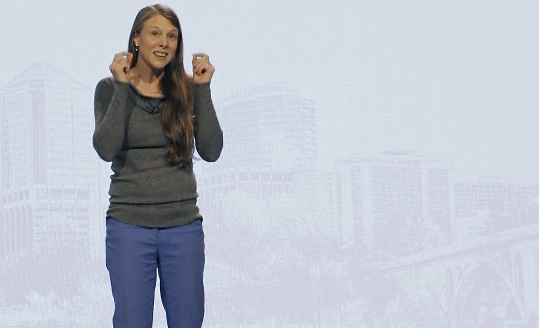 A woman with long brown hair, wearing a long-sleeved gray top and blue pants, speaking on stage in front of a light backdrop with faint cityscape outline.